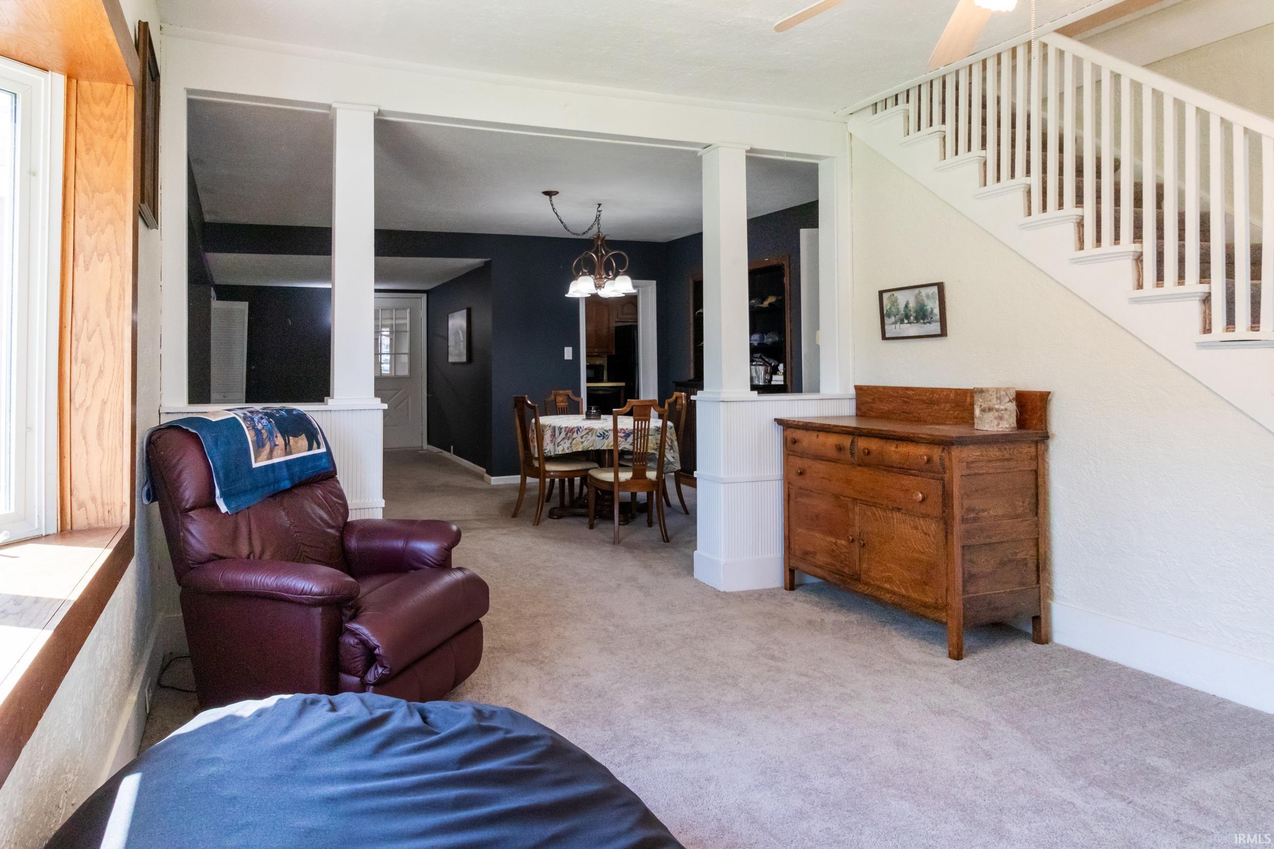 Carpeted living area featuring a prominent staircase with white balusters, a wooden window seat, and an open entryway with white columns