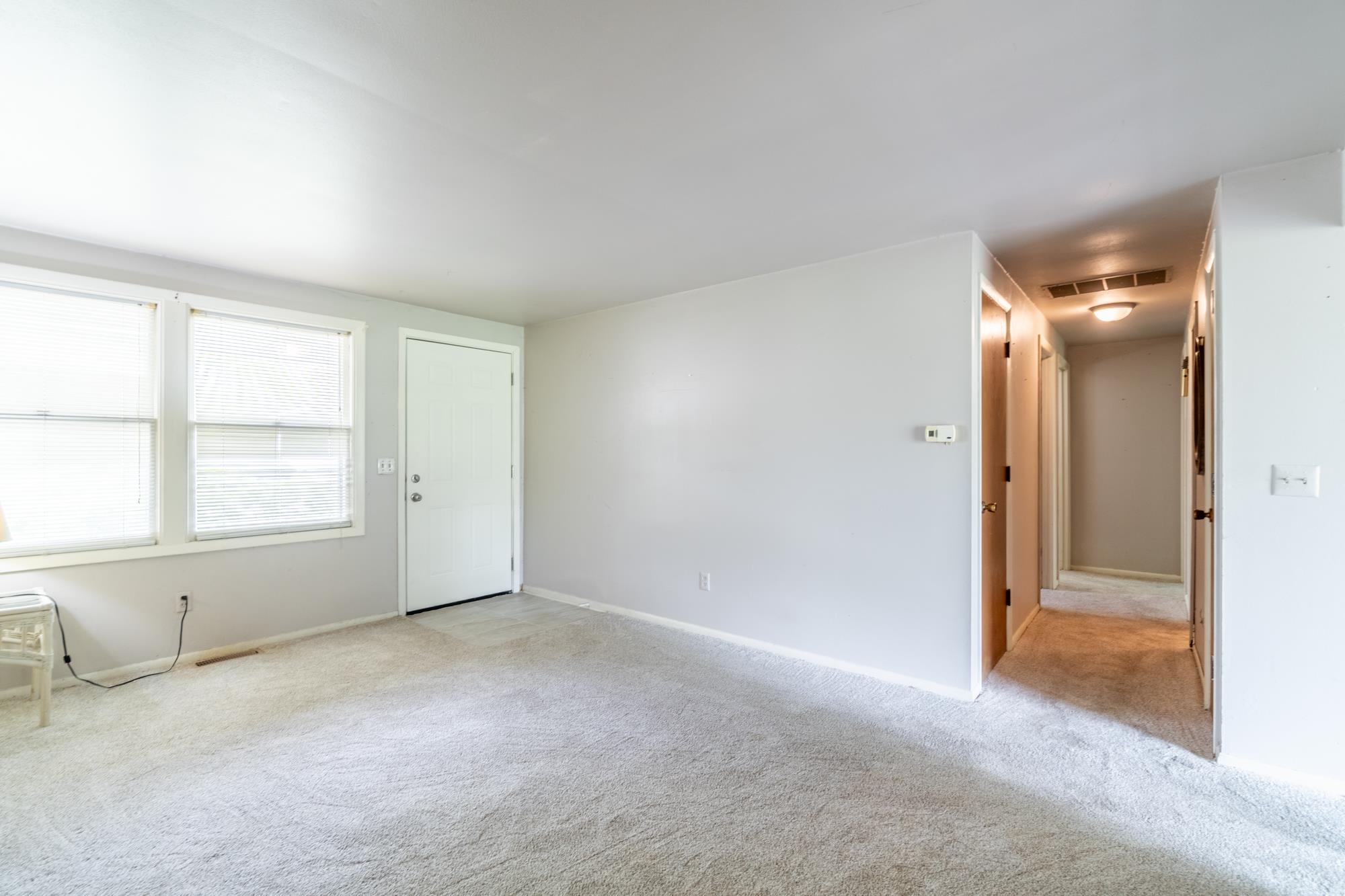 Spacious room featuring light-colored carpet flooring, a white paneled entry door, two windows with blinds, light gray wall paint, and a hallway with additional doors