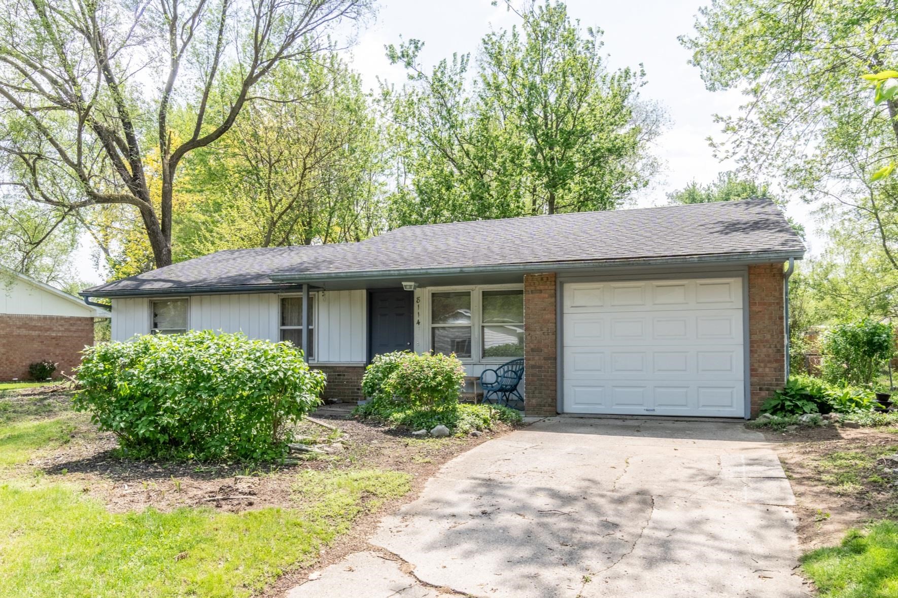 Ranch-style exterior with brick and siding facade, featuring a white paneled garage door, a dark asphalt shingle roof, and mature perimeter trees