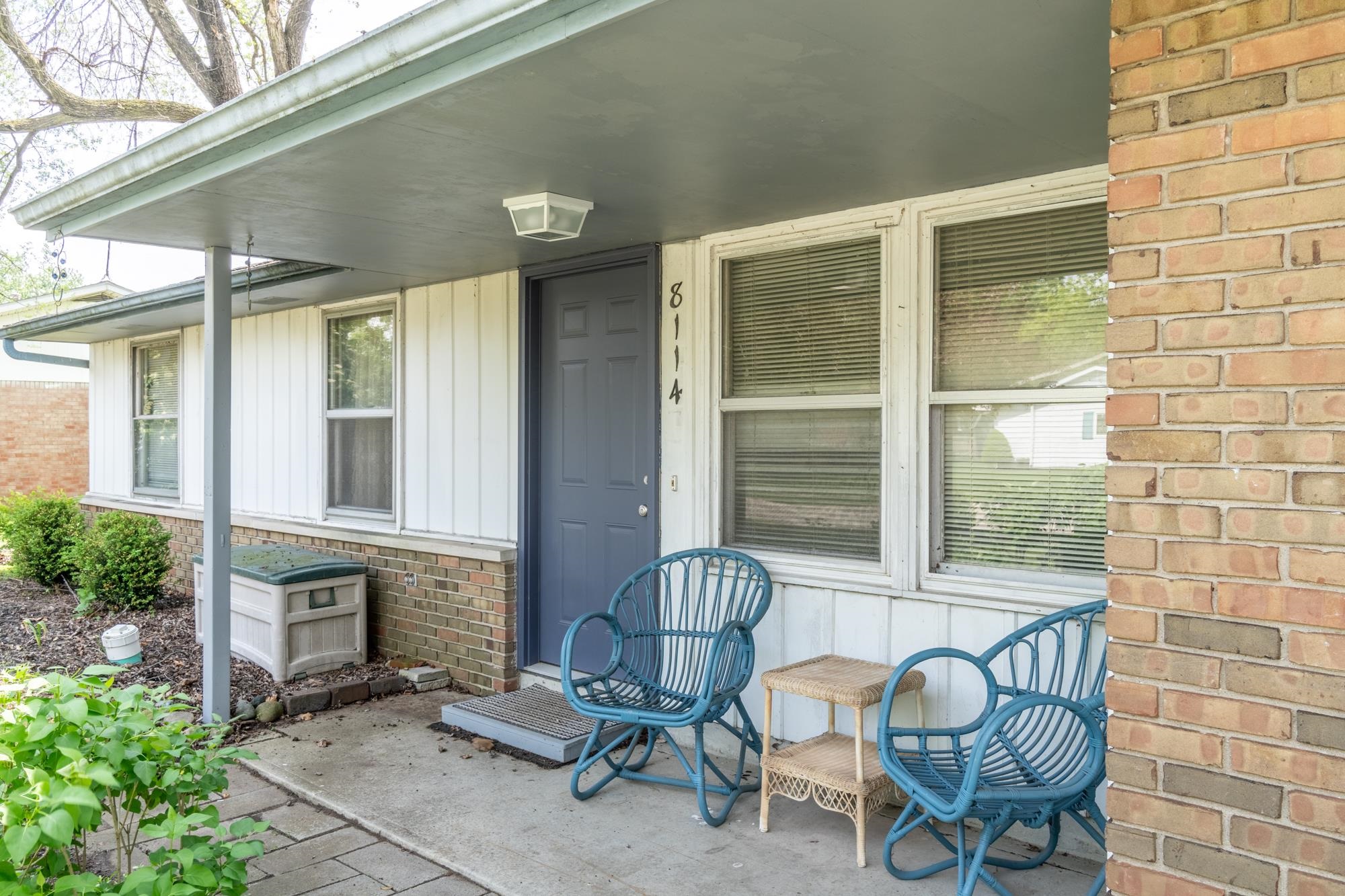 Covered front porch featuring a dark blue entry door, white vertical siding, and a brick facade
