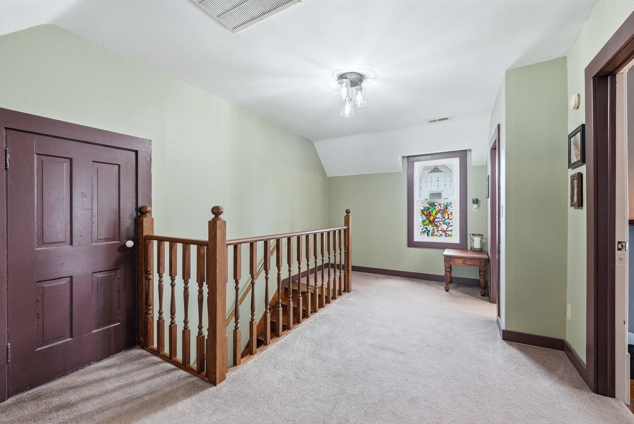 Carpeted hallway featuring a wood-finish staircase railing, paneled doors, and a decorative window