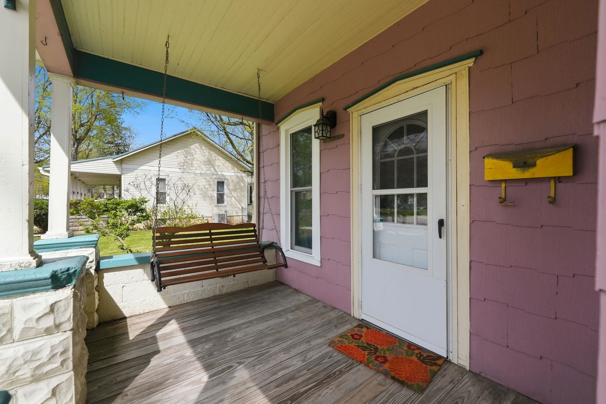 Covered front porch featuring wood-finish flooring, a white storm door with arched divided lites, and a dark wood-finish porch swing