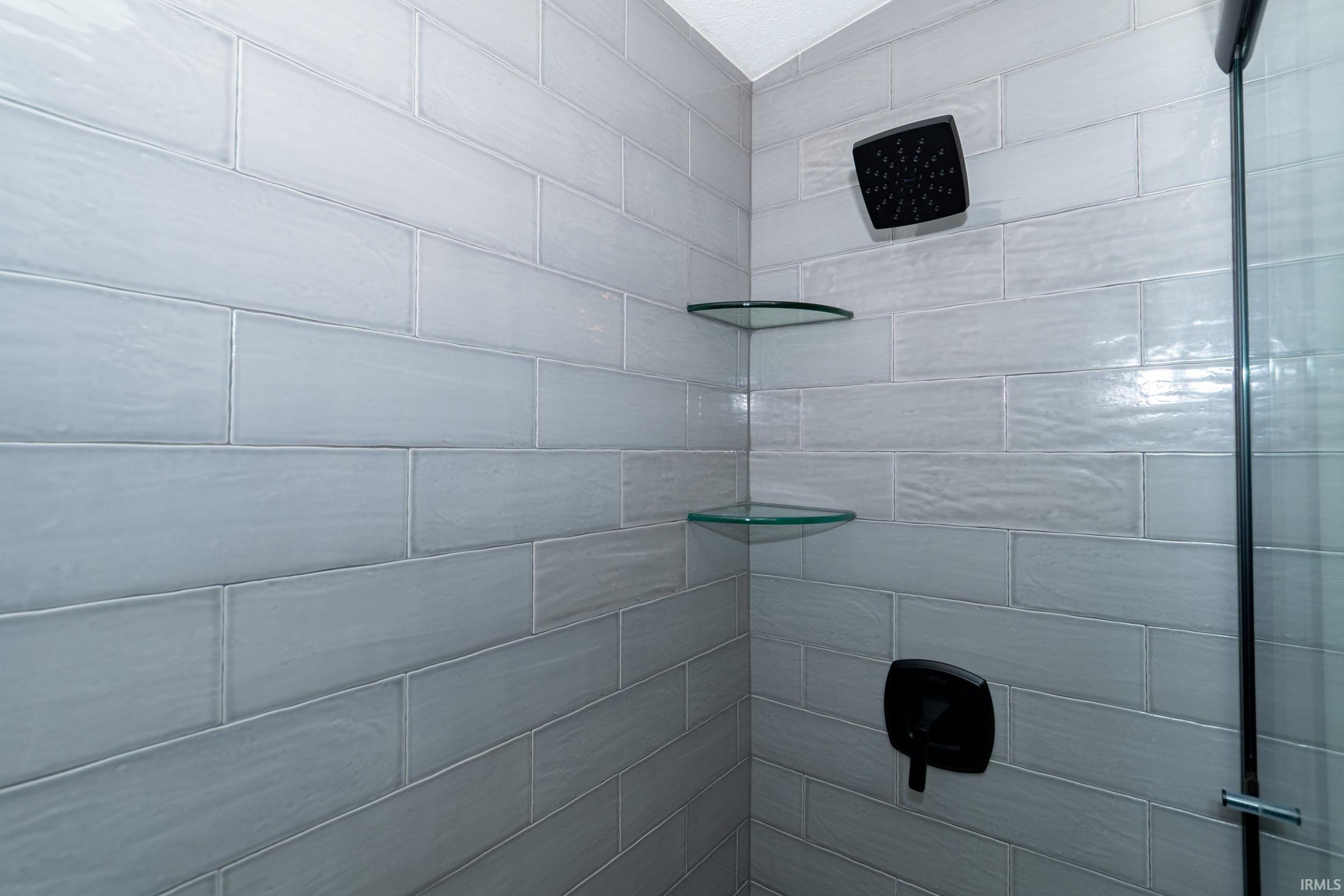 Shower enclosure featuring light-toned subway tile, a matte black rain shower head, coordinating matte black shower controls, two corner glass shelves, and a clear glass shower panel
