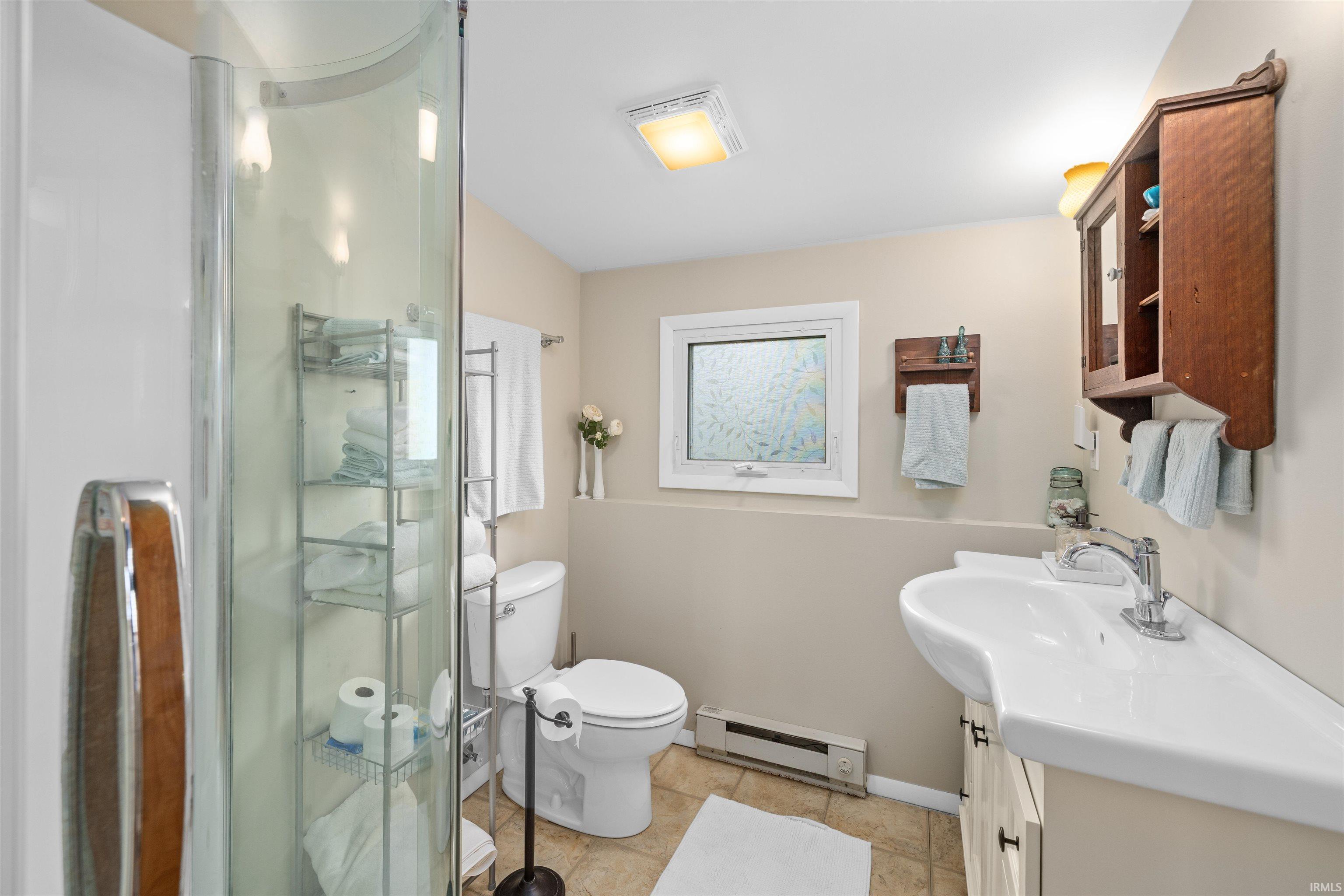 Bathroom featuring a curved glass shower enclosure, a ceramic vanity with a single basin, a toilet, a privacy window, and tiled flooring