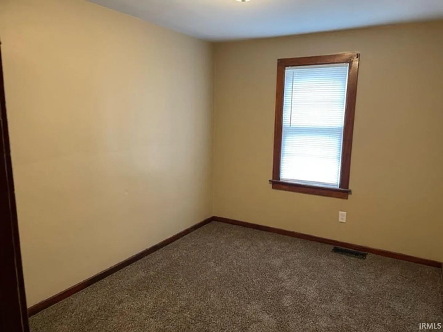 Interior room featuring neutral wall paint, rich wood trim around a window, and textured carpeting