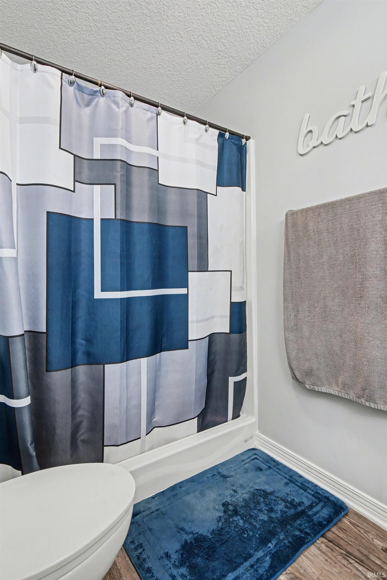 Bathtub with shower curtain, white toilet, and wood-finish flooring