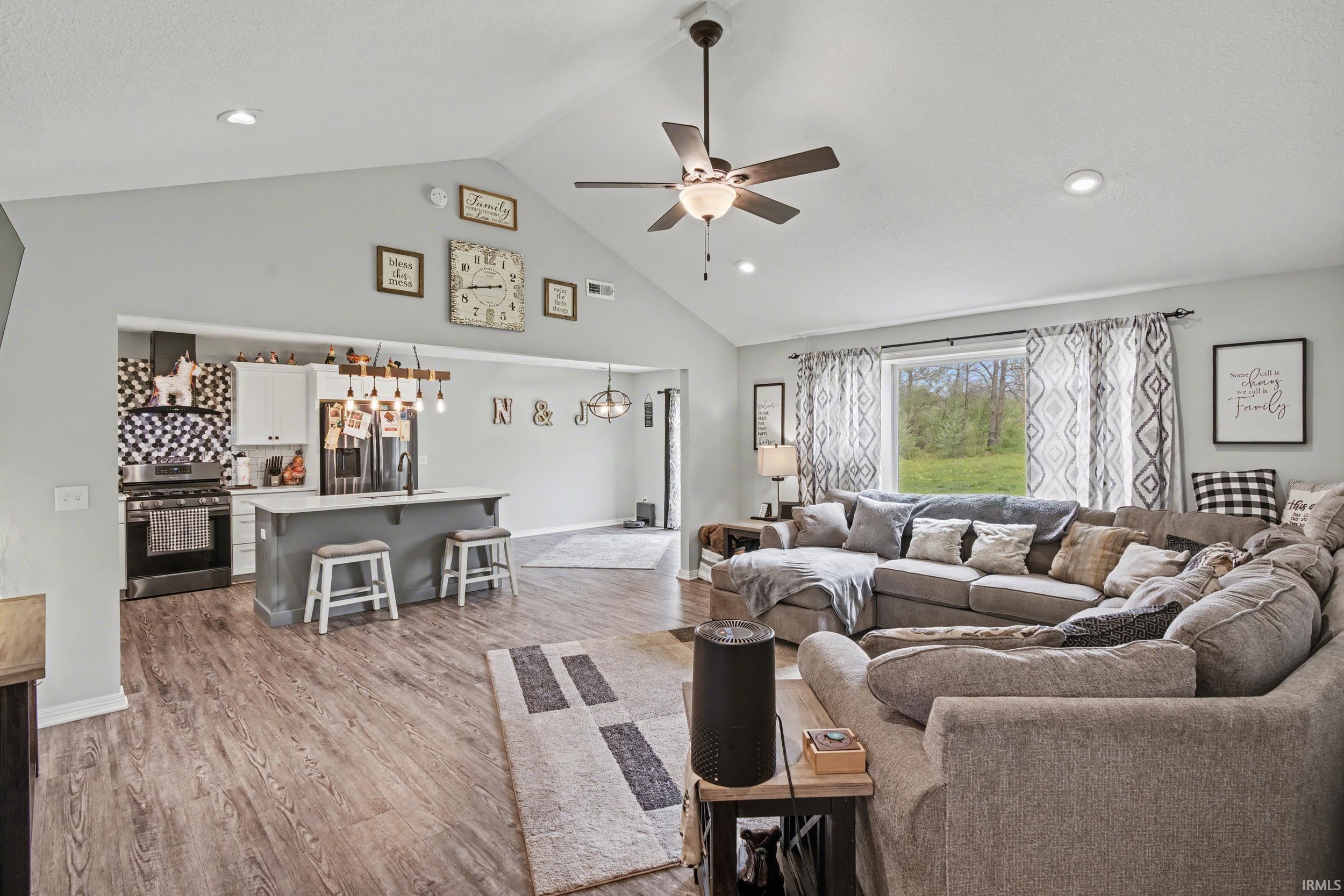 Open-concept living space featuring vaulted ceilings, wood-finish flooring, a kitchen island with seating, a built-in range hood, and a ceiling fan with integrated lighting