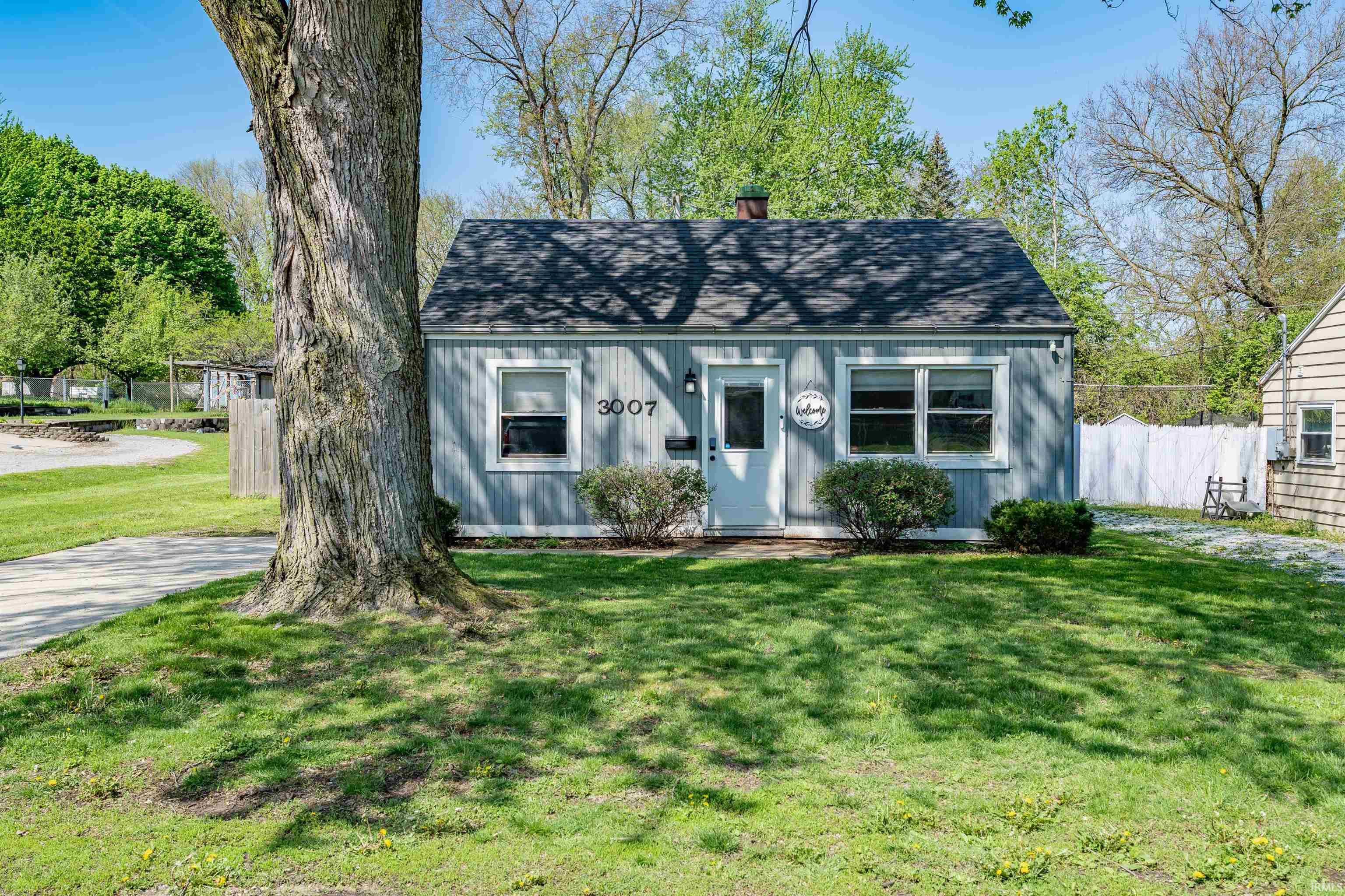 Light blue exterior siding with white trim, a dark shingle roof, and a mature tree providing natural shade
