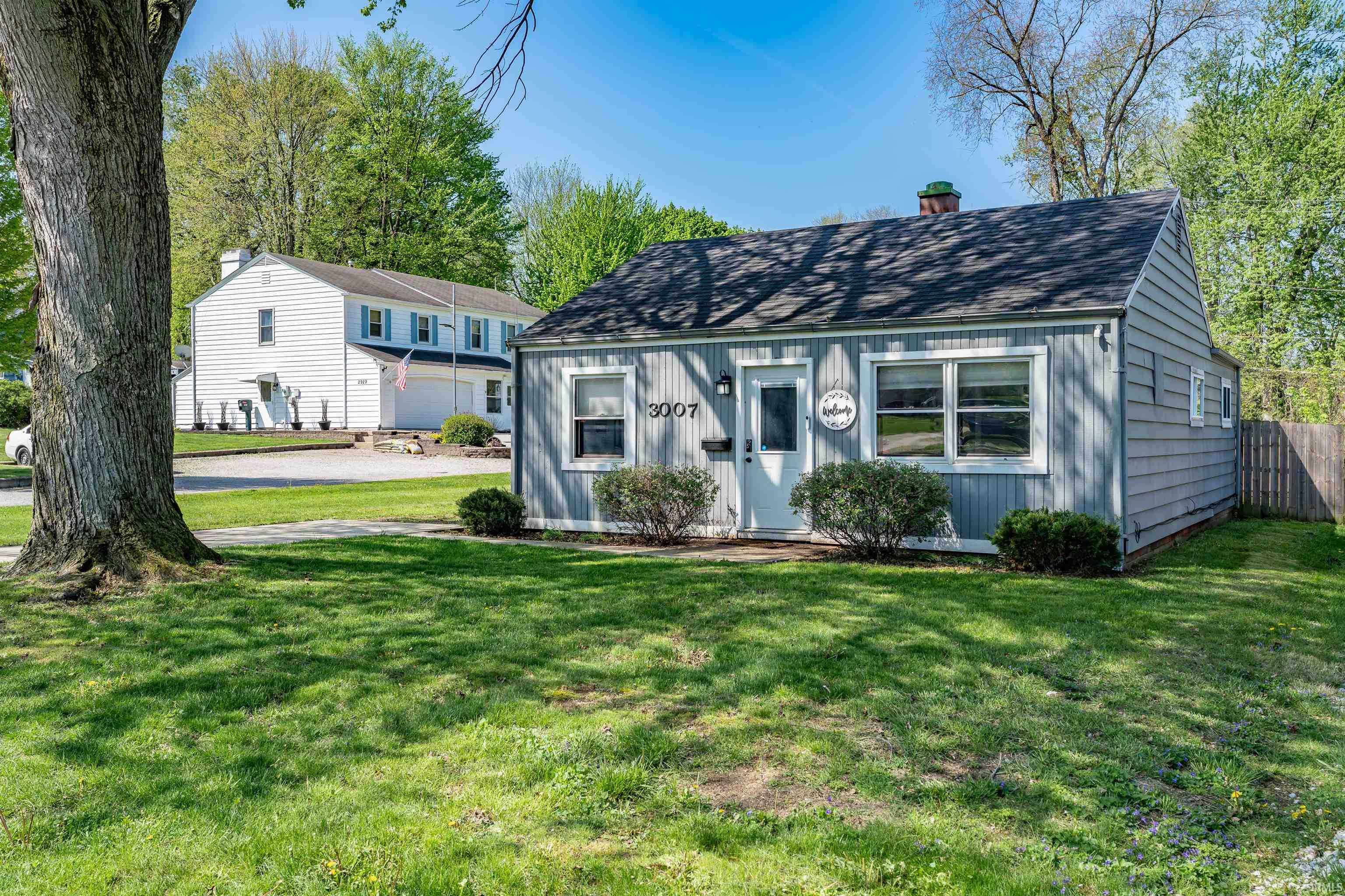 Gray siding exterior featuring a white front door, multiple windows with white trim, a dark shingle roof, and established front landscaping