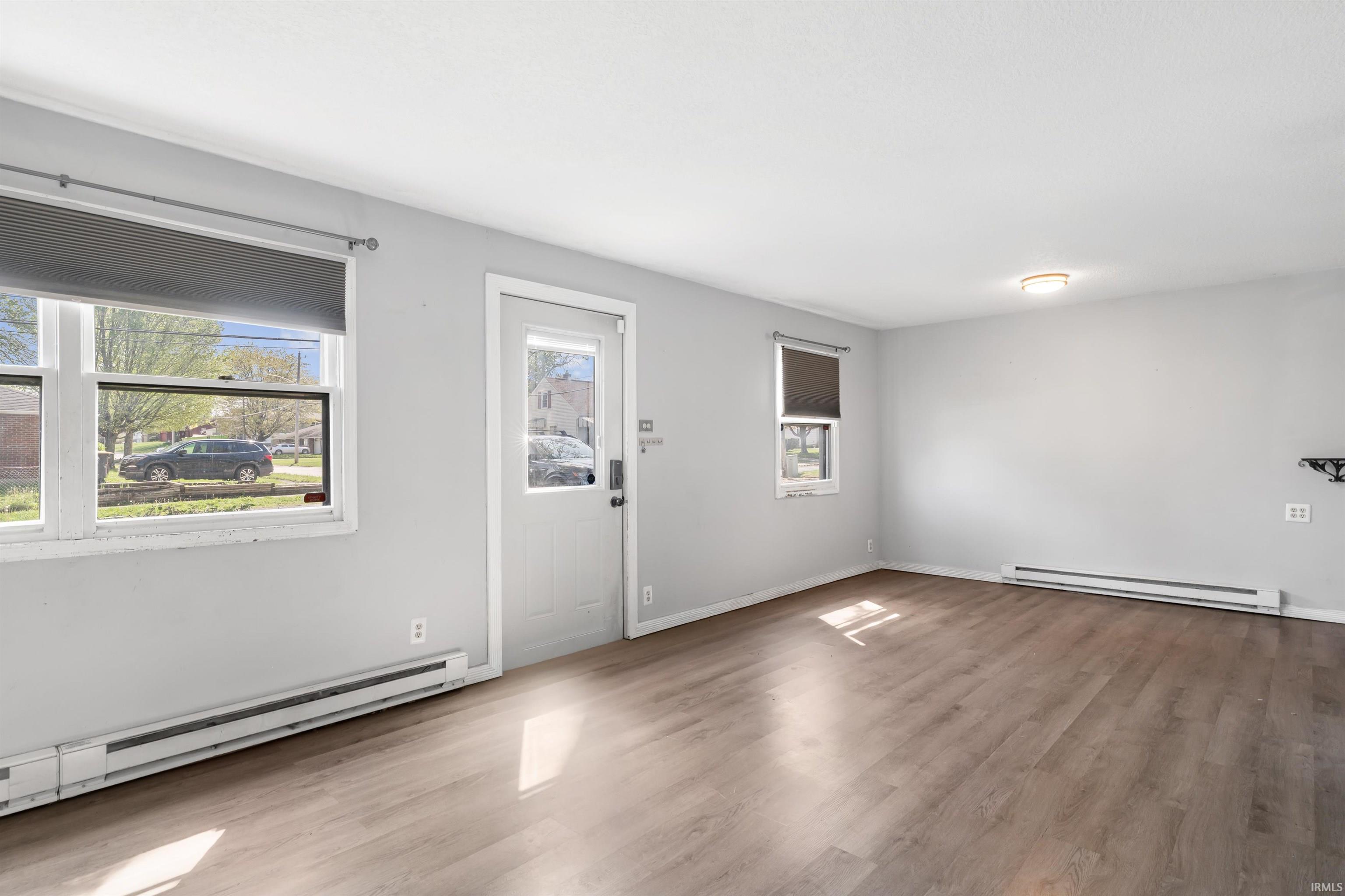 Living area featuring wood-finish flooring, light gray wall paint, white trim, multiple windows with roller shades, and a ceiling-mounted light fixture