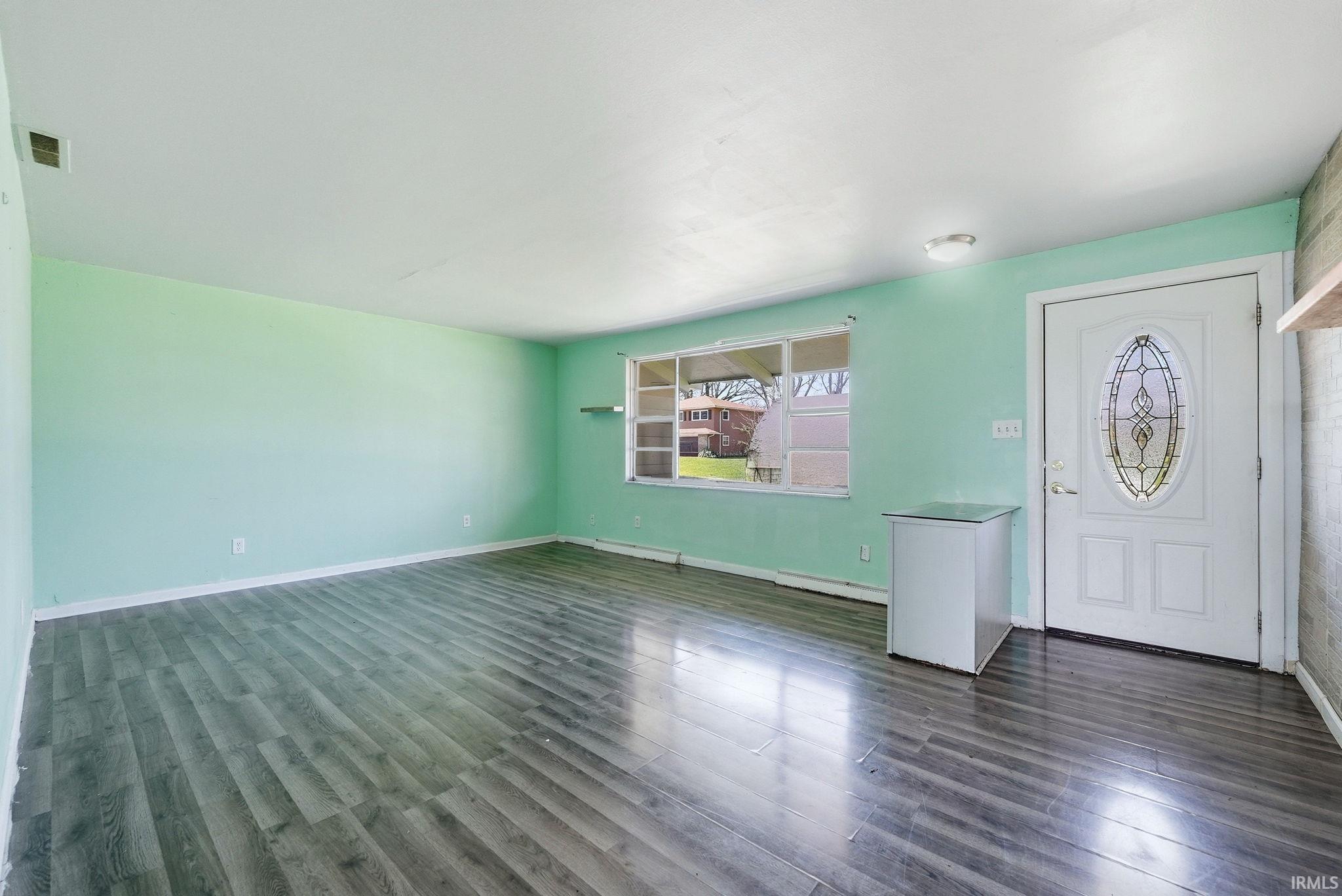 Spacious room featuring wood-finish flooring, a white entry door with an oval stained glass insert, and a large window providing natural illumination