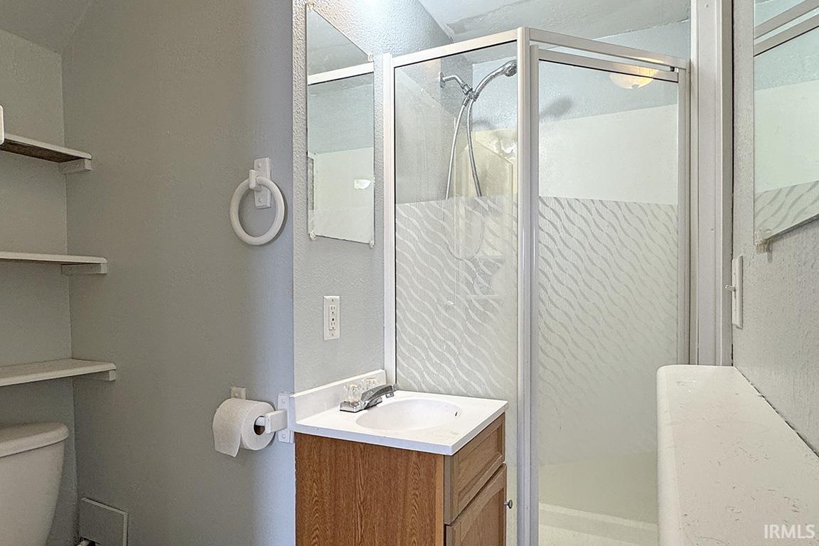 Compact bathroom featuring a stand-up shower with textured glass enclosure, a vanity with wood-finish cabinetry and a white countertop, and open shelving