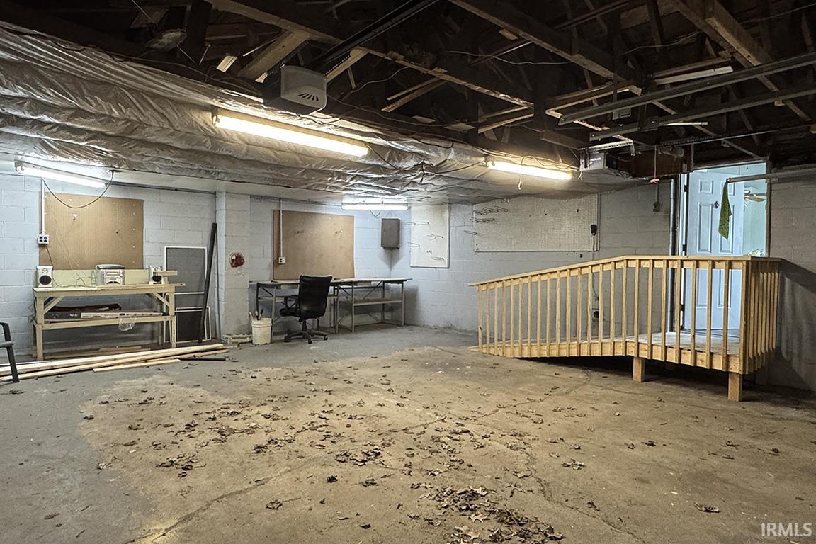Spacious utility area featuring exposed wood beam ceiling, concrete flooring, fluorescent lighting fixtures, and a wood ramp leading to an interior door