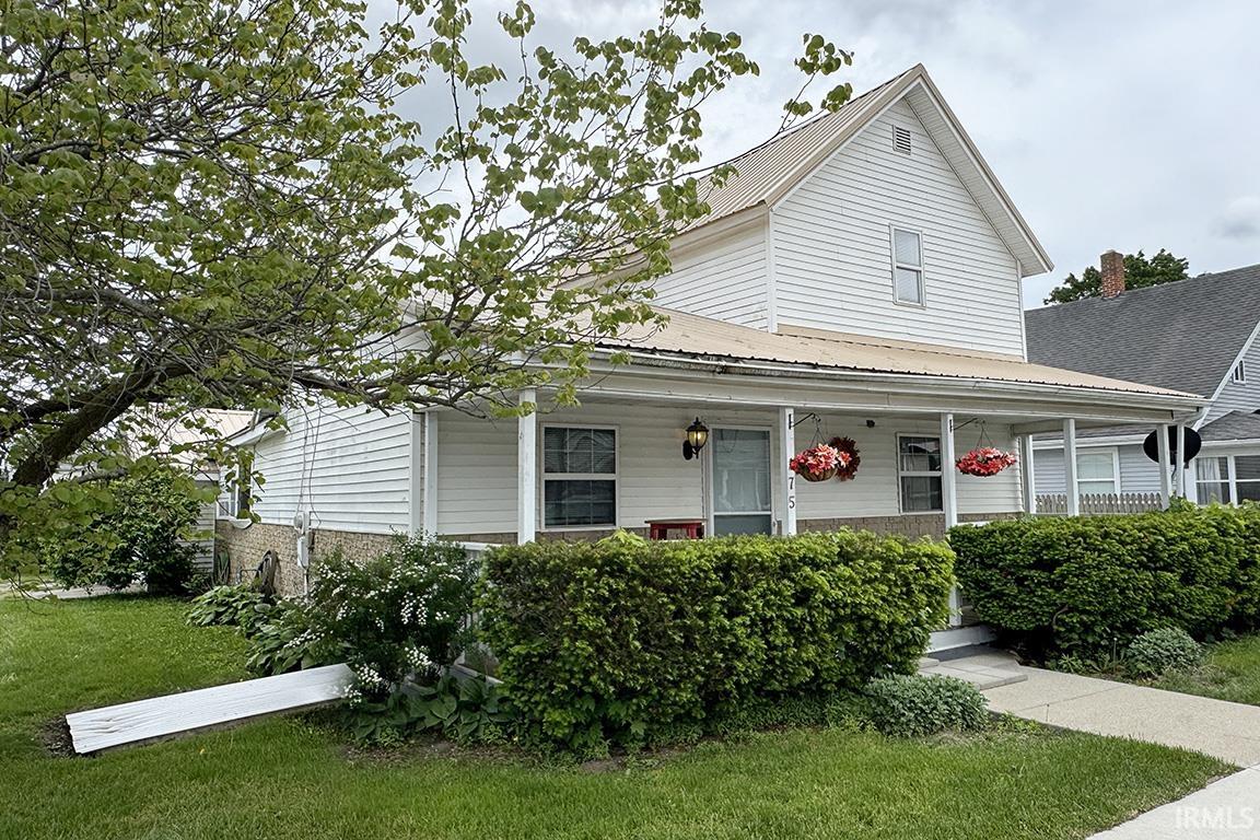 White siding exterior featuring a covered porch, established shrubbery, a paved walkway, and a metal roof