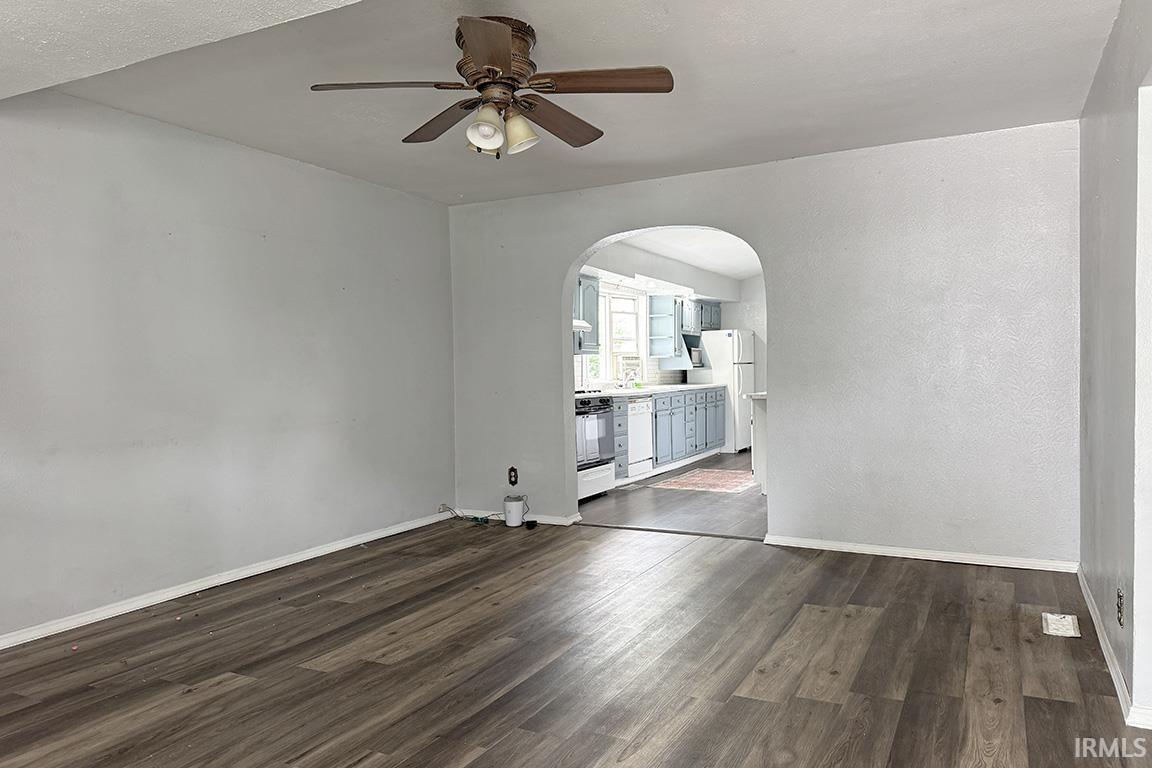 Spacious room featuring wood-finish flooring, a ceiling fan, and an arched doorway leading to a kitchen with light blue cabinetry, white countertops, and white appliances