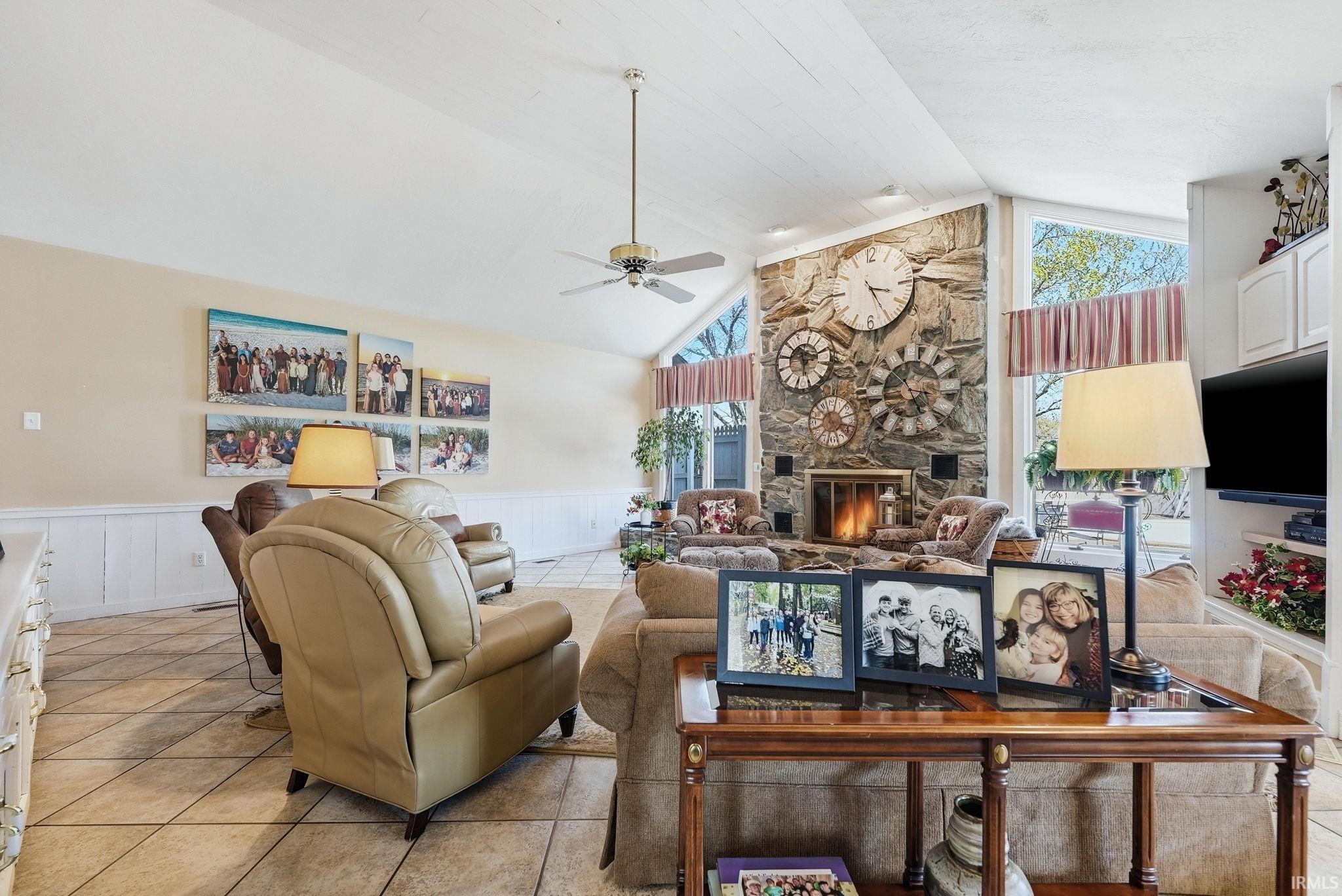 Living area featuring light tile patterned floors, wainscoting, a stone fireplace, a ceiling fan, and a decorative wall