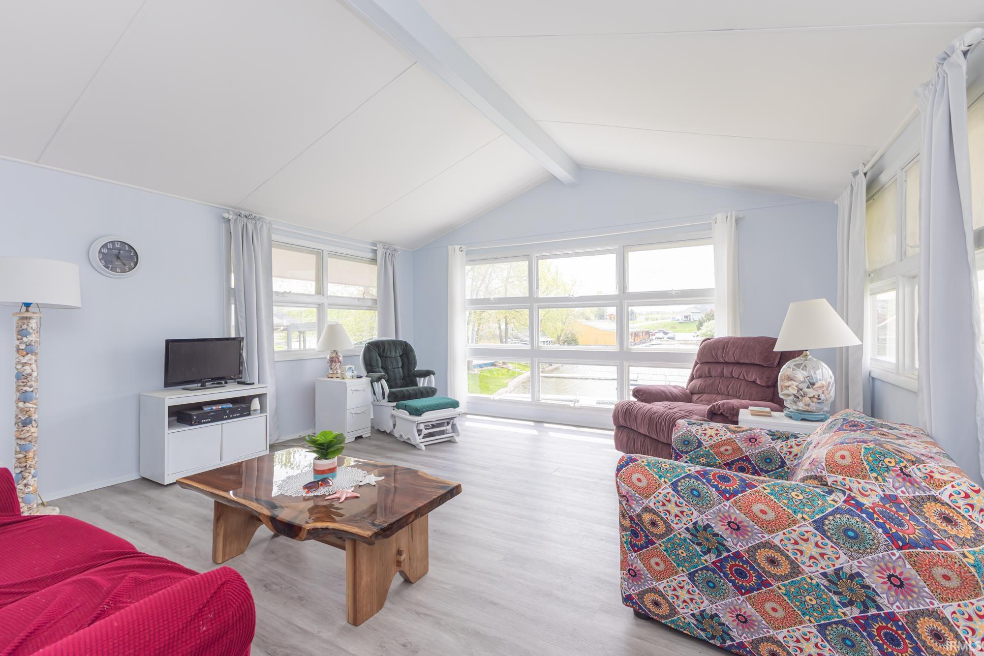 Bright living area featuring abundant natural light from multiple windows, light wood-finish flooring, and a vaulted ceiling with exposed beams