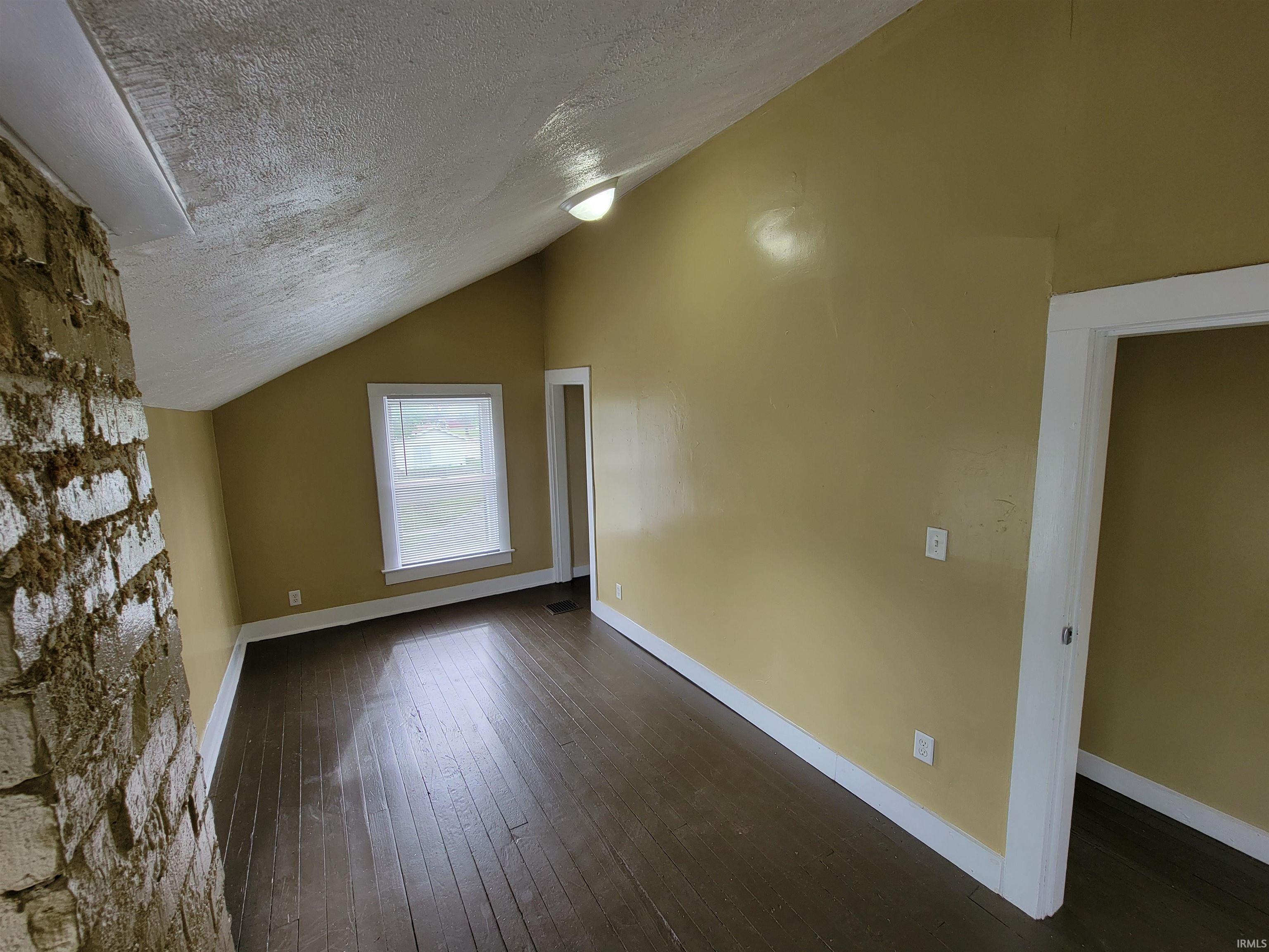 Room featuring a stone-clad architectural element, angled ceiling, wood-finish flooring, and a window with white trim