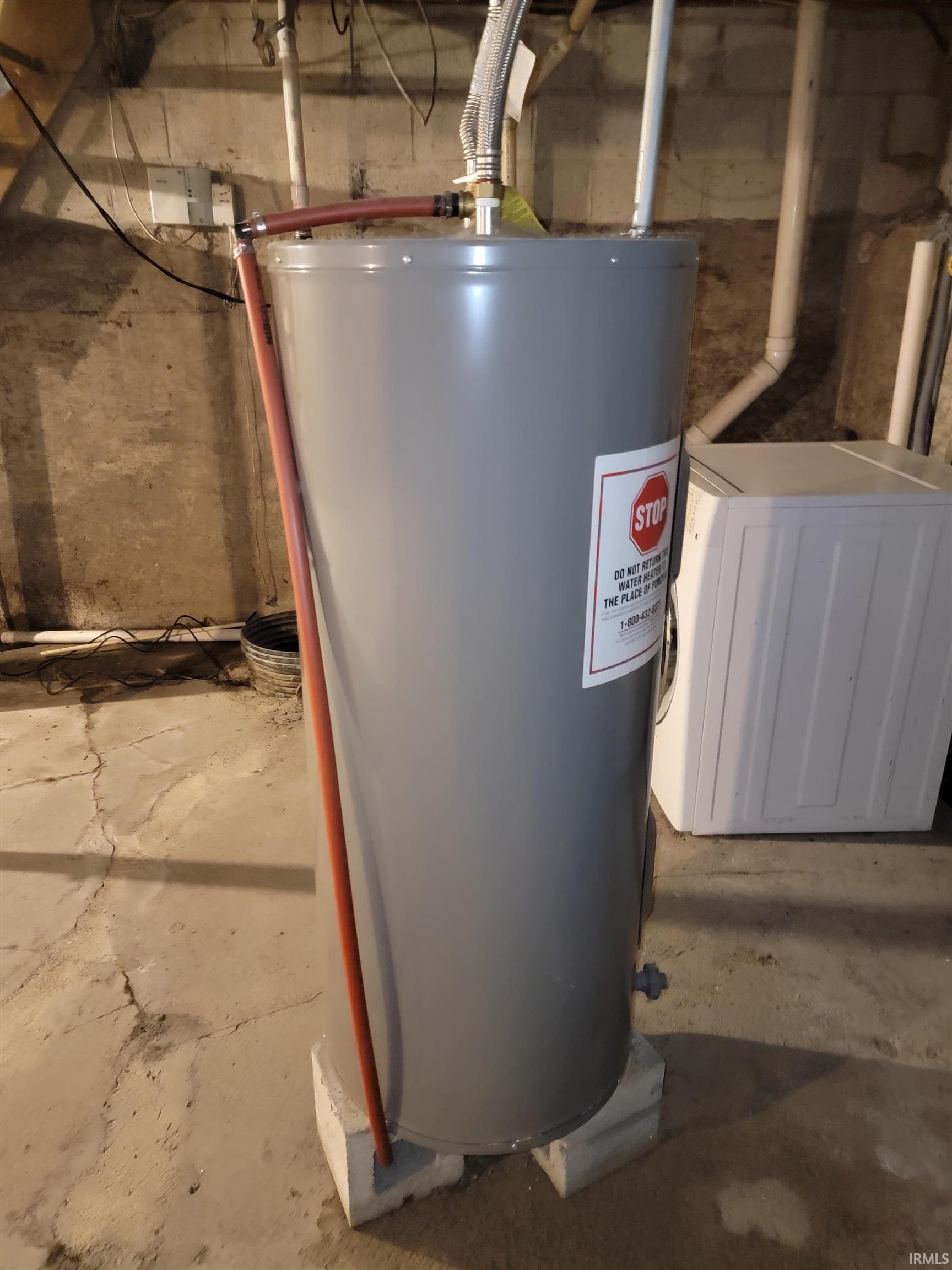 Gray water heater on elevated concrete blocks, featuring visible plumbing connections and a concrete floor
