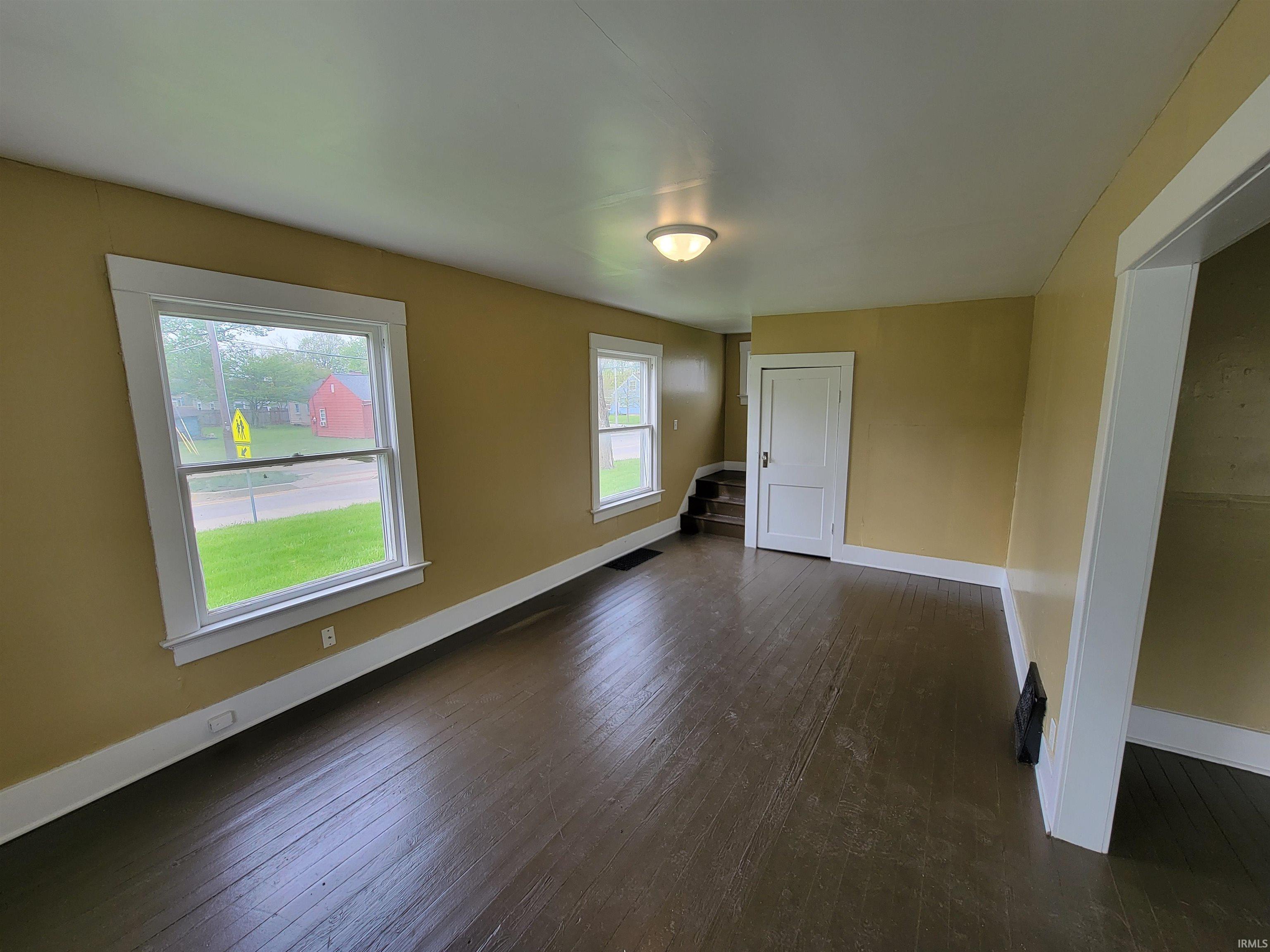 Spacious room featuring dark wood-finish flooring, light-colored walls, white trim, and two large windows