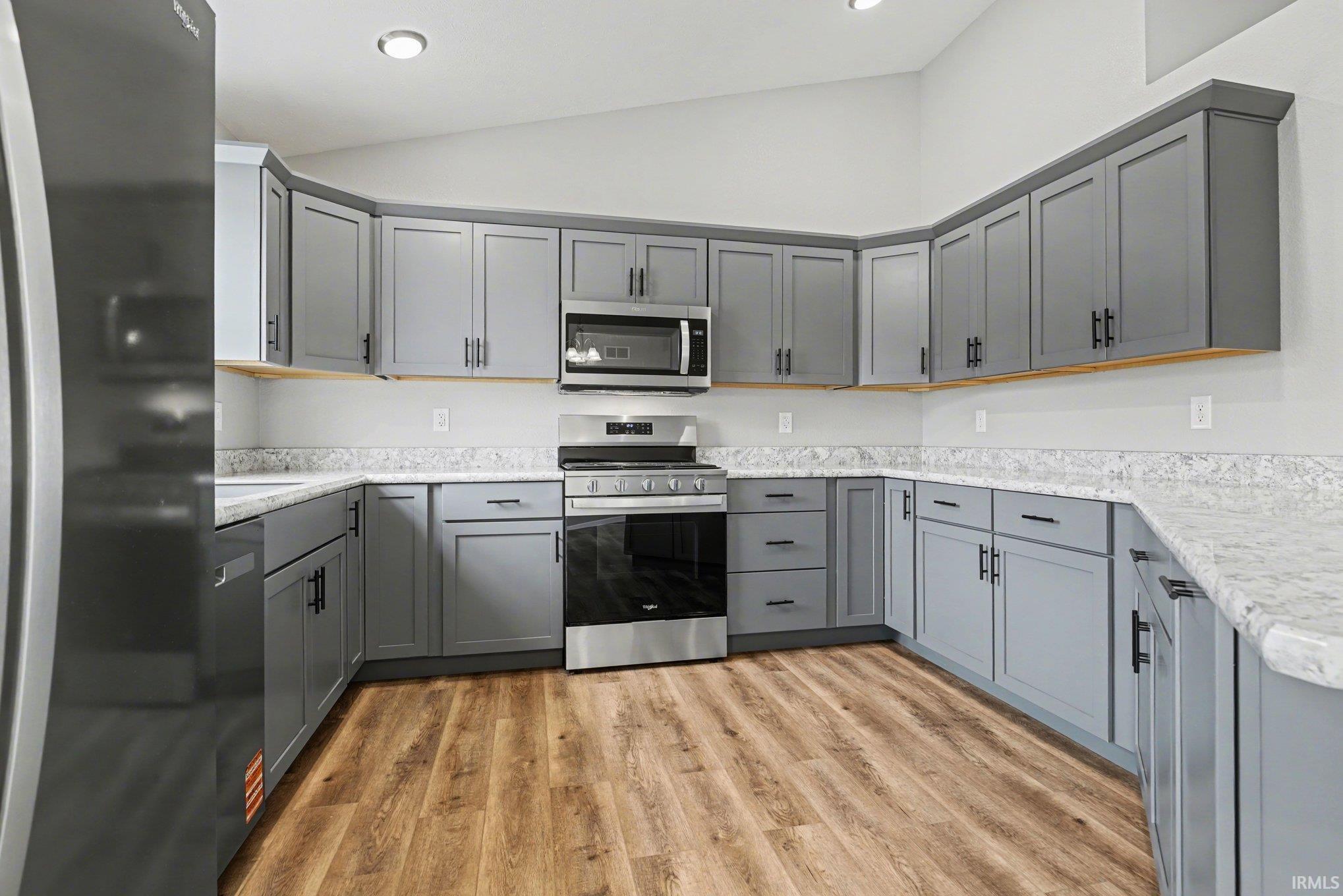 Expansive kitchen featuring wood-finish flooring, light-toned countertops, gray cabinetry with black hardware, stainless steel appliances, and recessed lighting