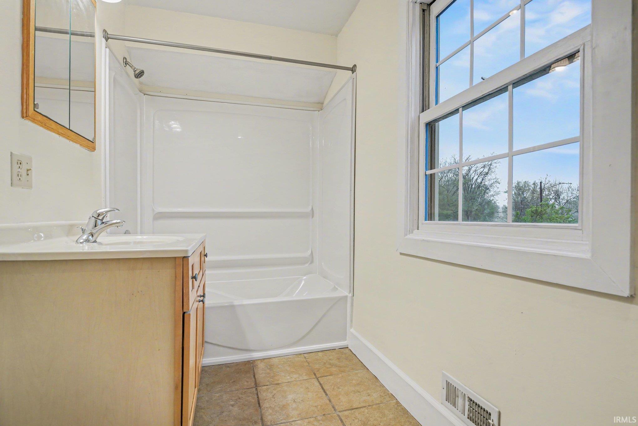 Bathroom with a single-pane window, white tub and shower surround, wood-finish vanity with a single sink, and neutral-toned tile flooring