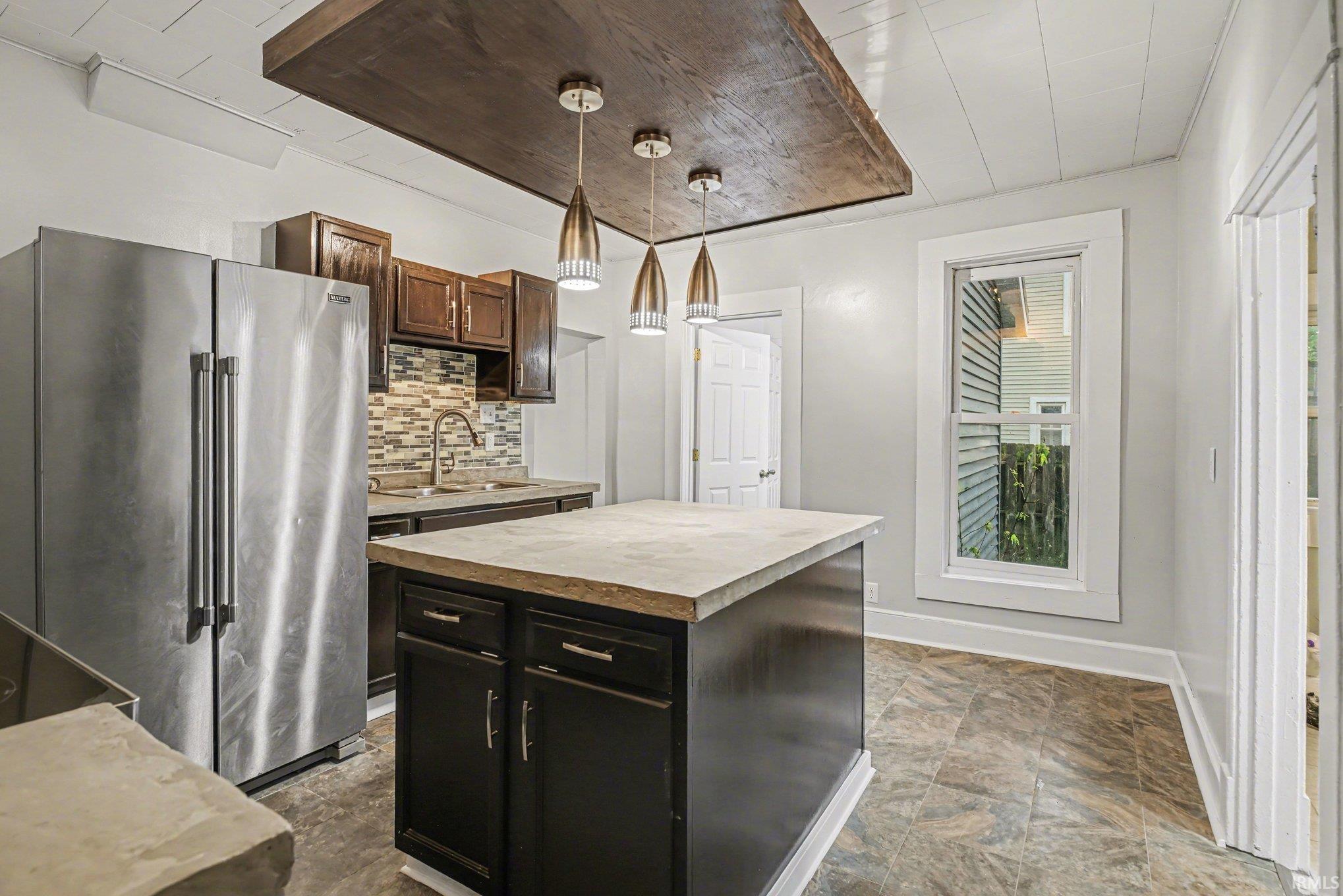 Kitchen featuring a central island with a light-toned countertop, dark wood cabinetry, a mosaic tile backsplash, and overhead pendant lighting