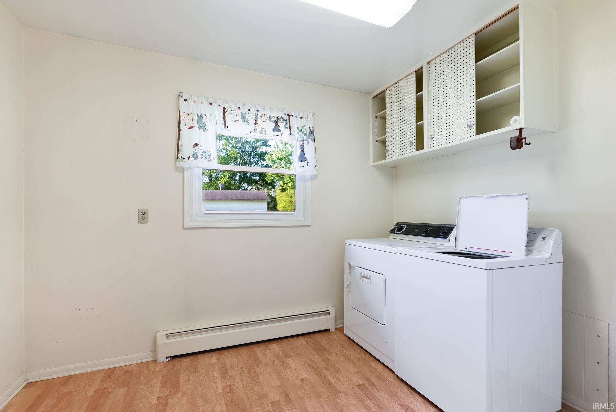 Dedicated laundry area featuring wood-finish flooring, a white window frame, baseboard heating, and built-in upper cabinetry with sliding doors