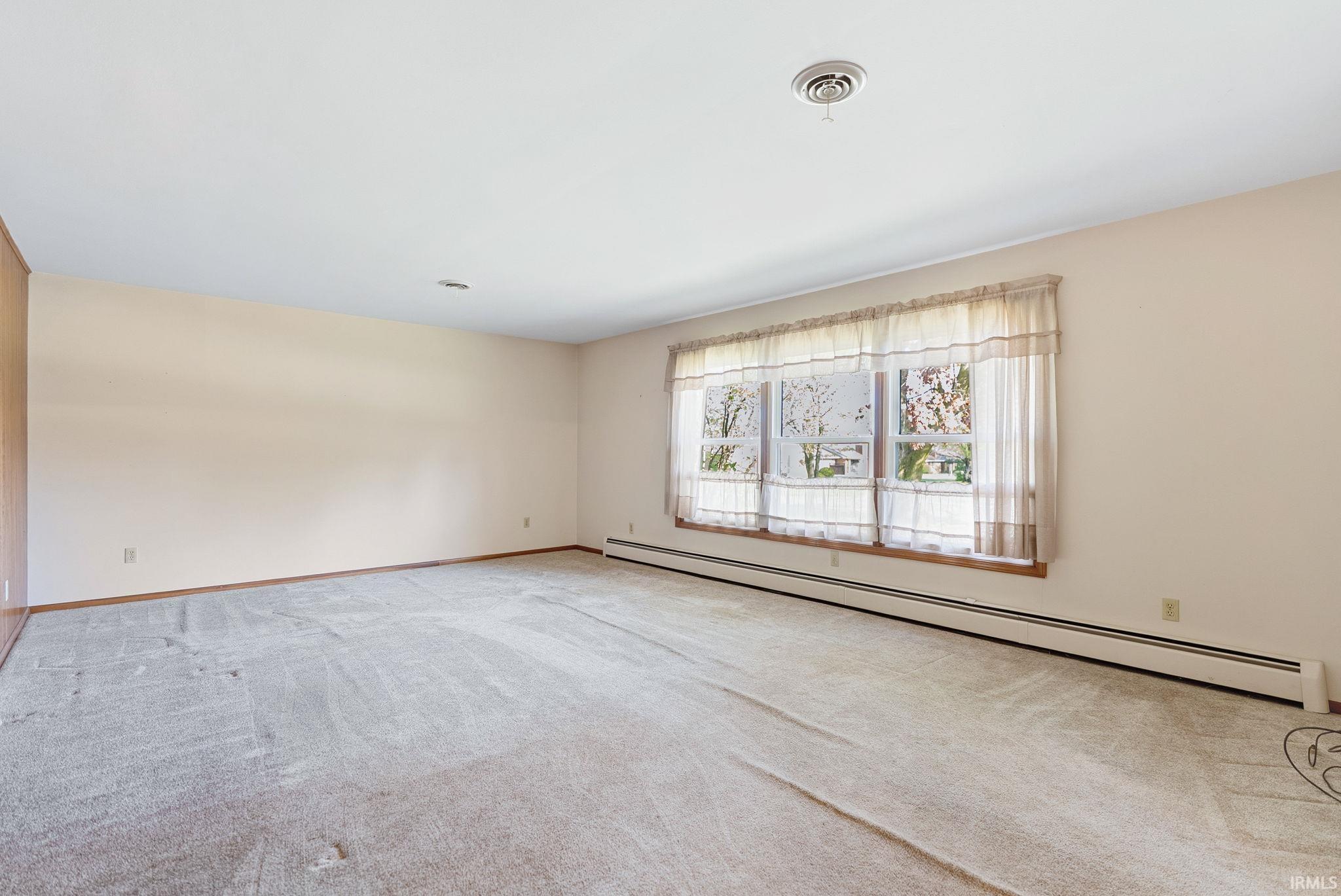 Spacious room featuring light-colored carpeting, neutral wall tones, and baseboard heating