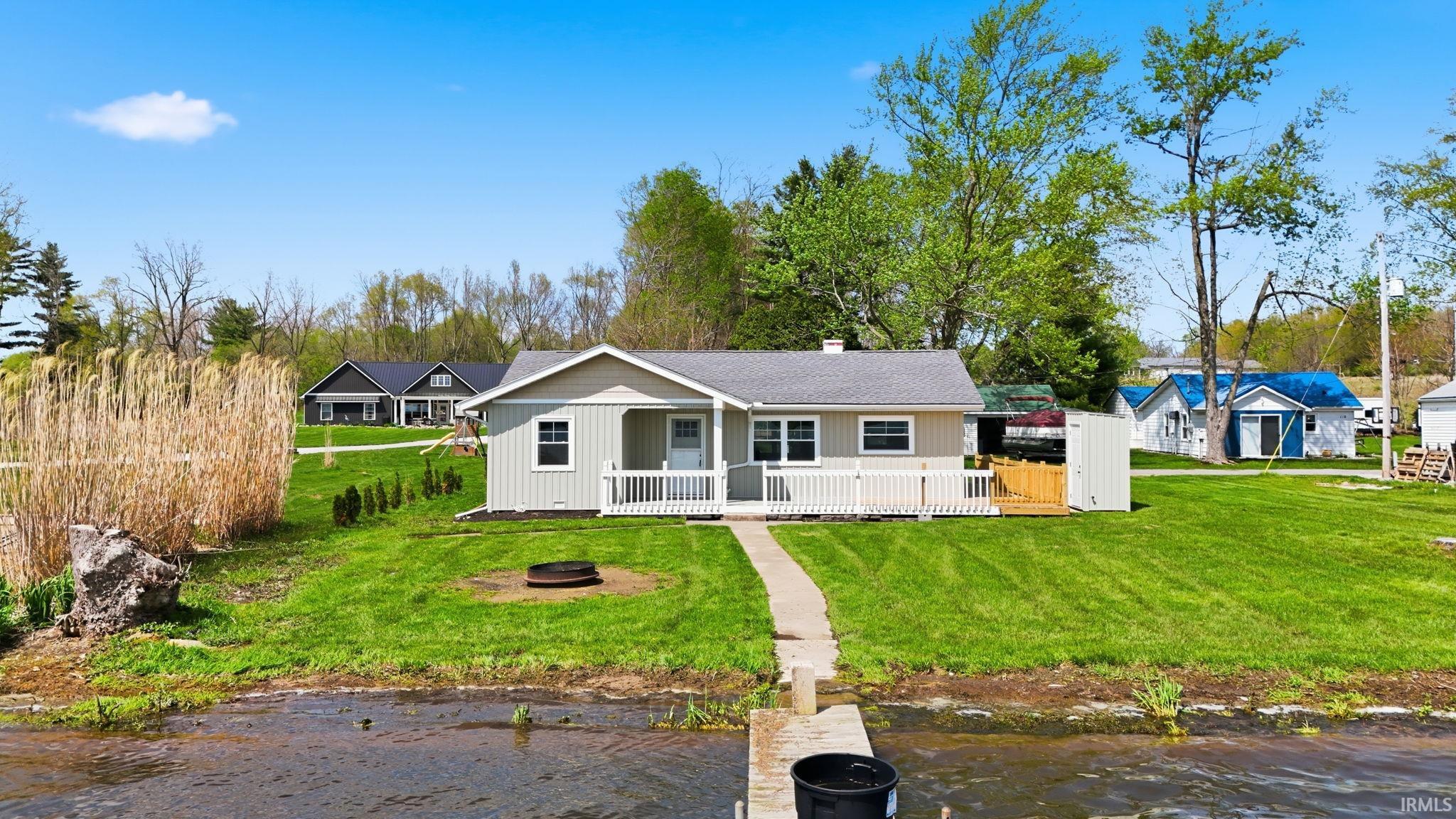 Waterfront property featuring a dock, a light-colored siding exterior, a front porch with white railings, a concrete walkway, and a spacious green lawn