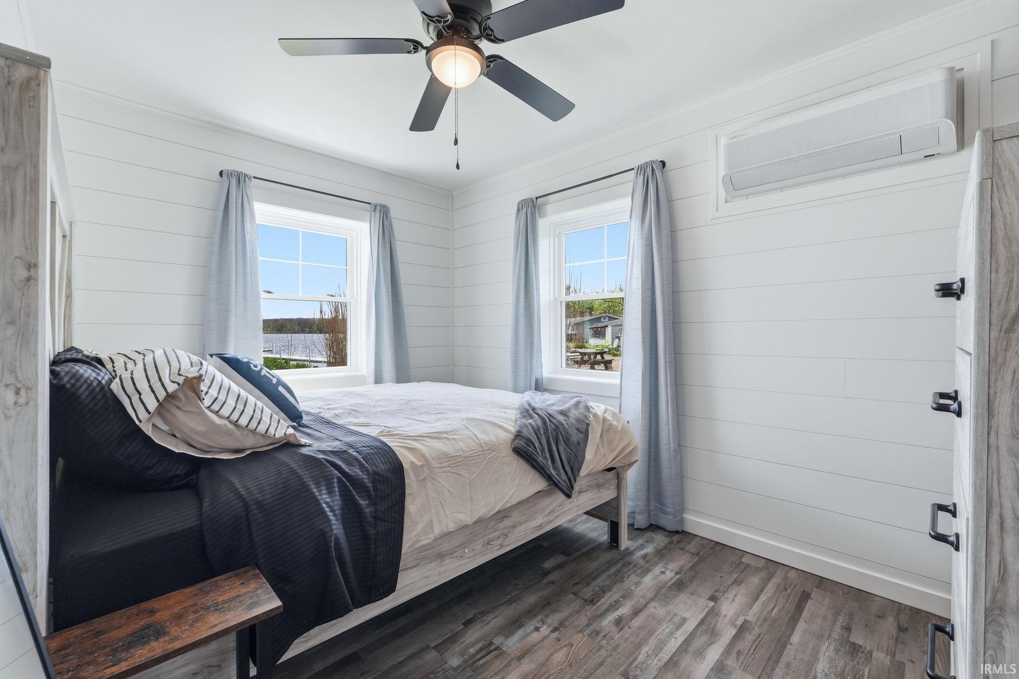 Bedroom featuring shiplap walls, wood-finish flooring, two windows, a ceiling fan, and a wall-mounted AC unit