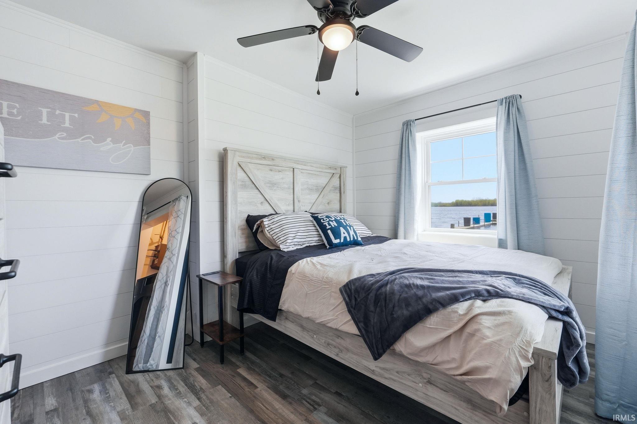 Waterfront bedroom featuring shiplap walls, wood-finish flooring, a ceiling fan with integrated lighting, and a window overlooking the water