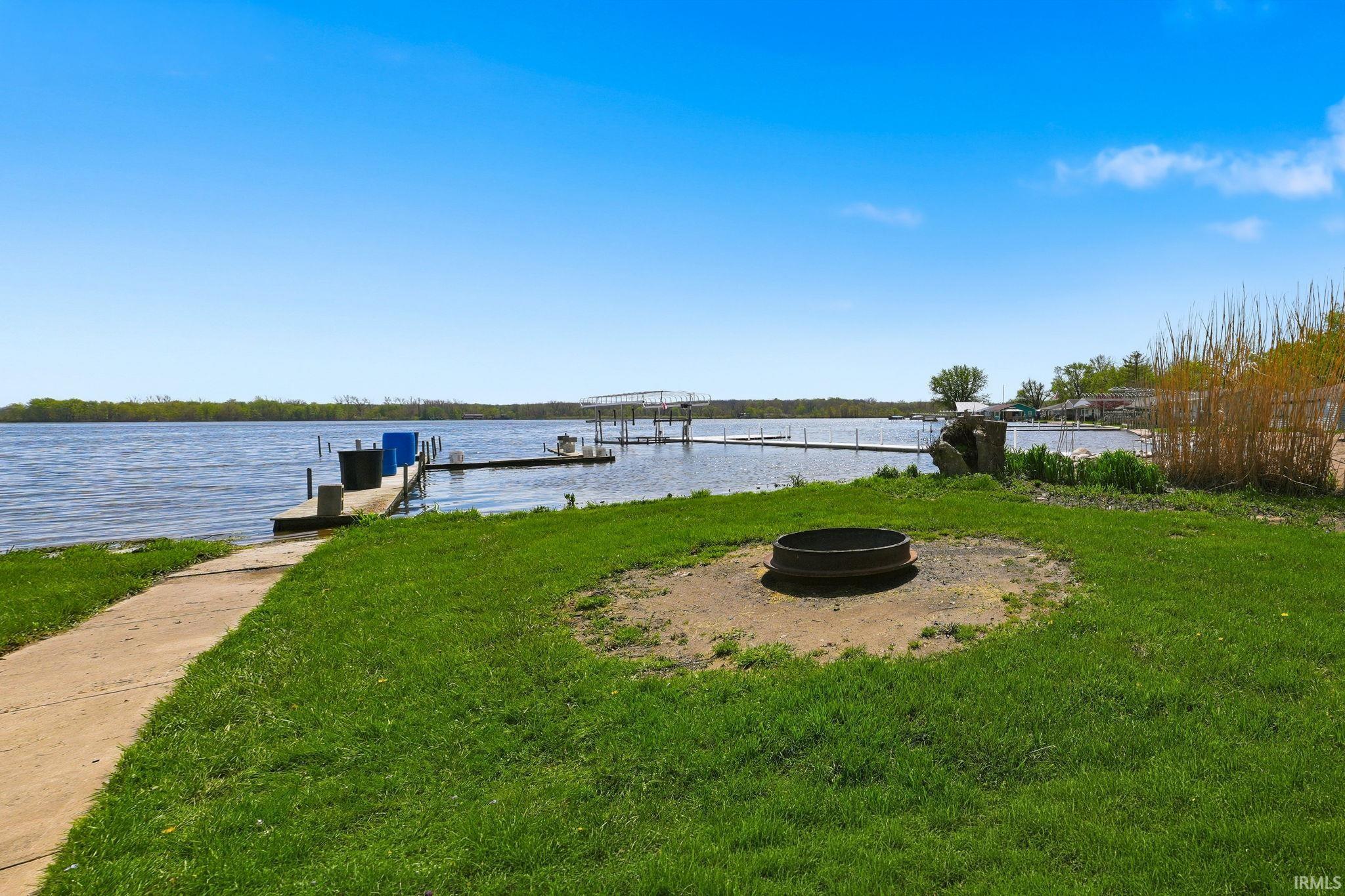 Waterfront property featuring a private dock, expansive water frontage, a concrete pathway, a built-in fire pit, and an open lawn area