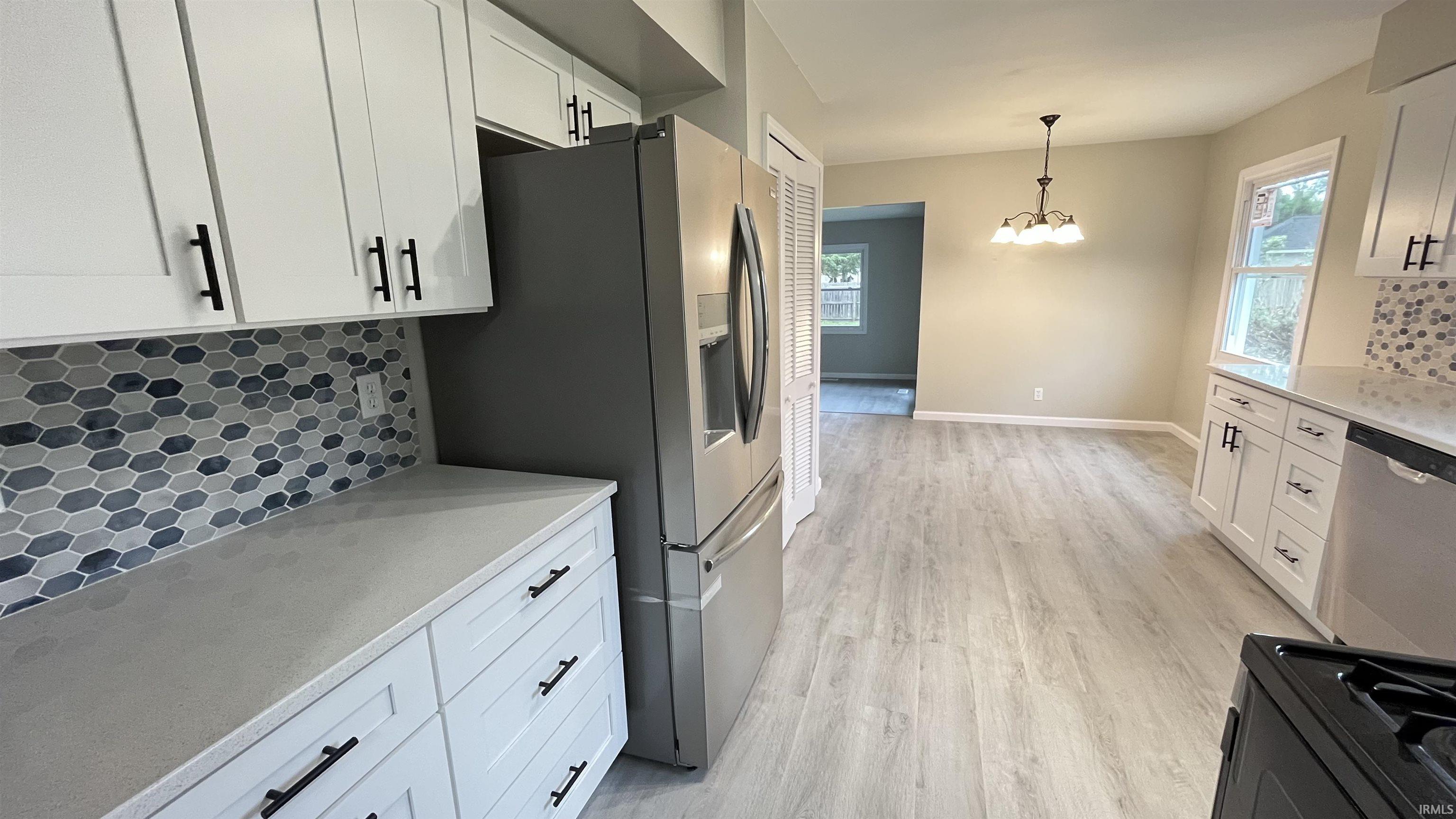 Modern kitchen featuring white cabinetry with matte black hardware, light-toned countertops, a hexagonal tile backsplash, stainless steel appliances, and light wood-finish flooring