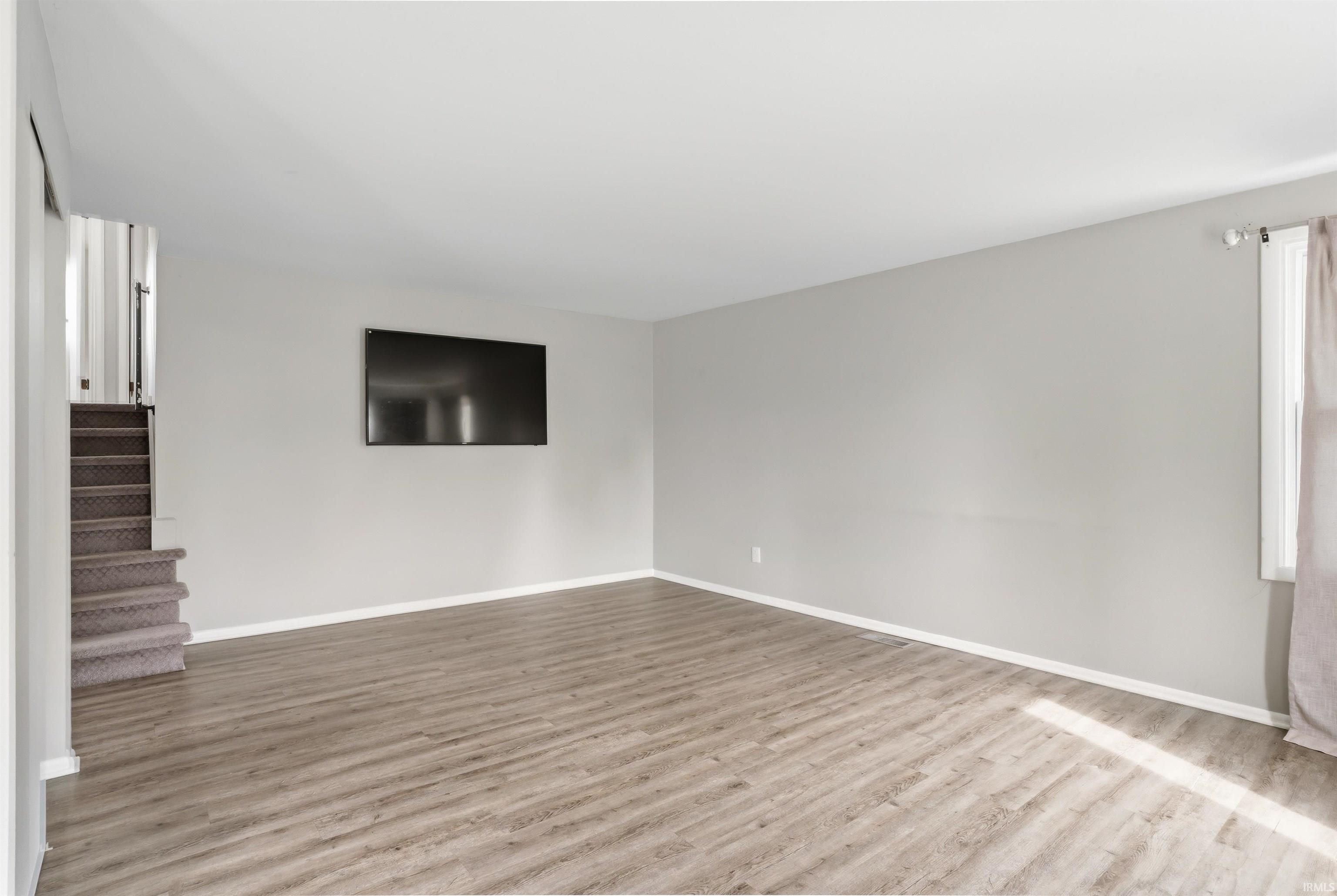 Spacious room featuring wood-finish flooring, light grey wall paint, white baseboards, and a window with natural light