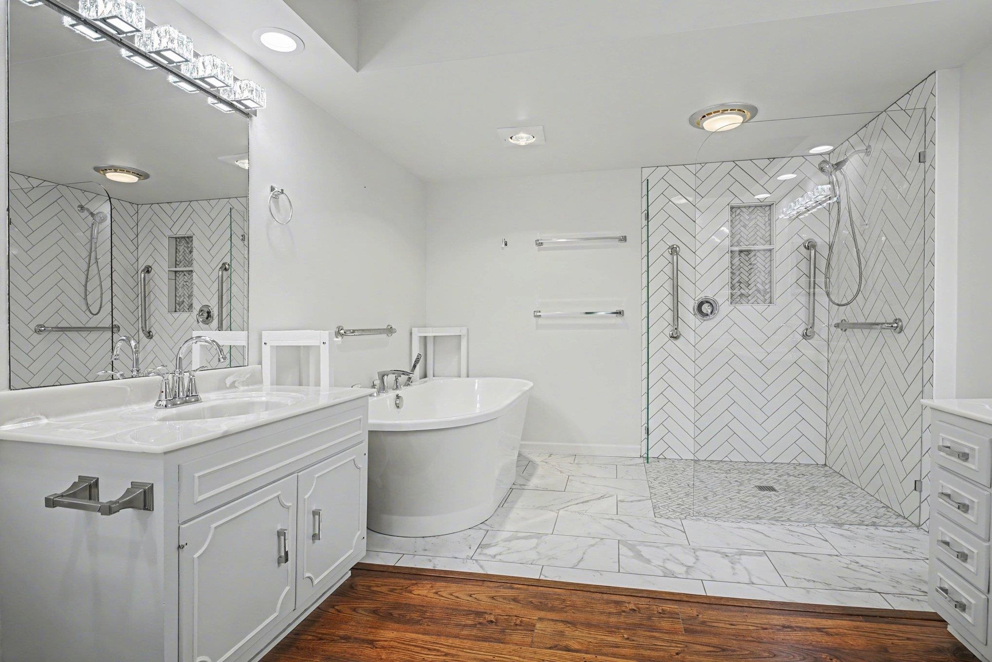 Bathroom featuring a spacious walk-in shower with herringbone tile, a freestanding soaking tub, dual white vanities with integrated sinks, and polished chrome fixtures