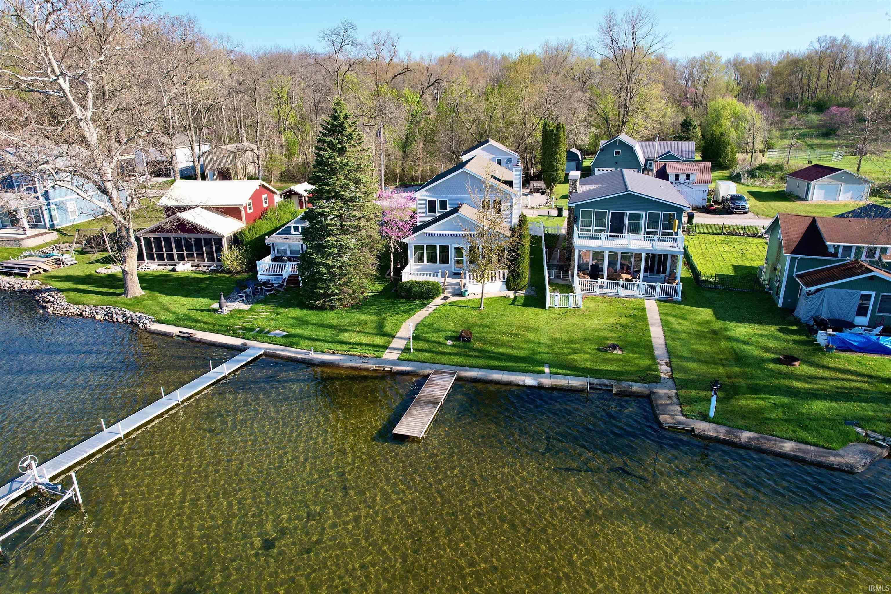Lakefront property featuring a private dock, lush green lawn, mature trees, and a concrete pathway leading to the water's edge