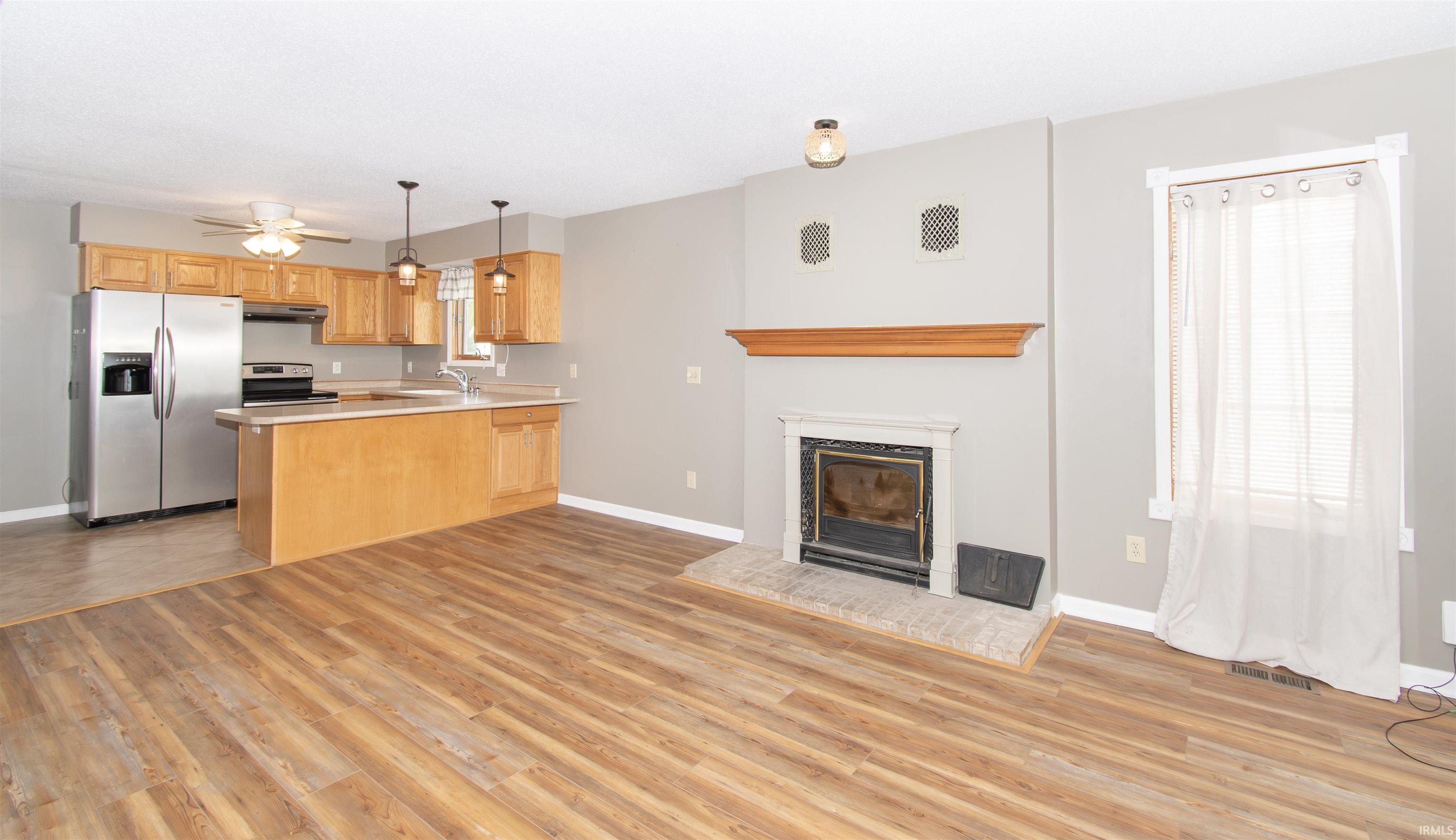 Open-concept living space featuring wood-finish flooring, a fireplace with a white mantel and hearth, and a kitchen with oak cabinetry and stainless steel appliances