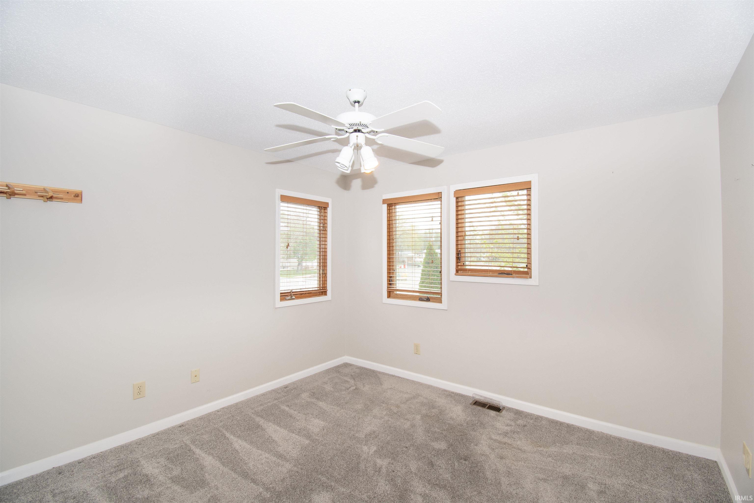 Neutral-toned room featuring a ceiling fan with light fixture, three windows with wood-finish blinds, and gray carpeting