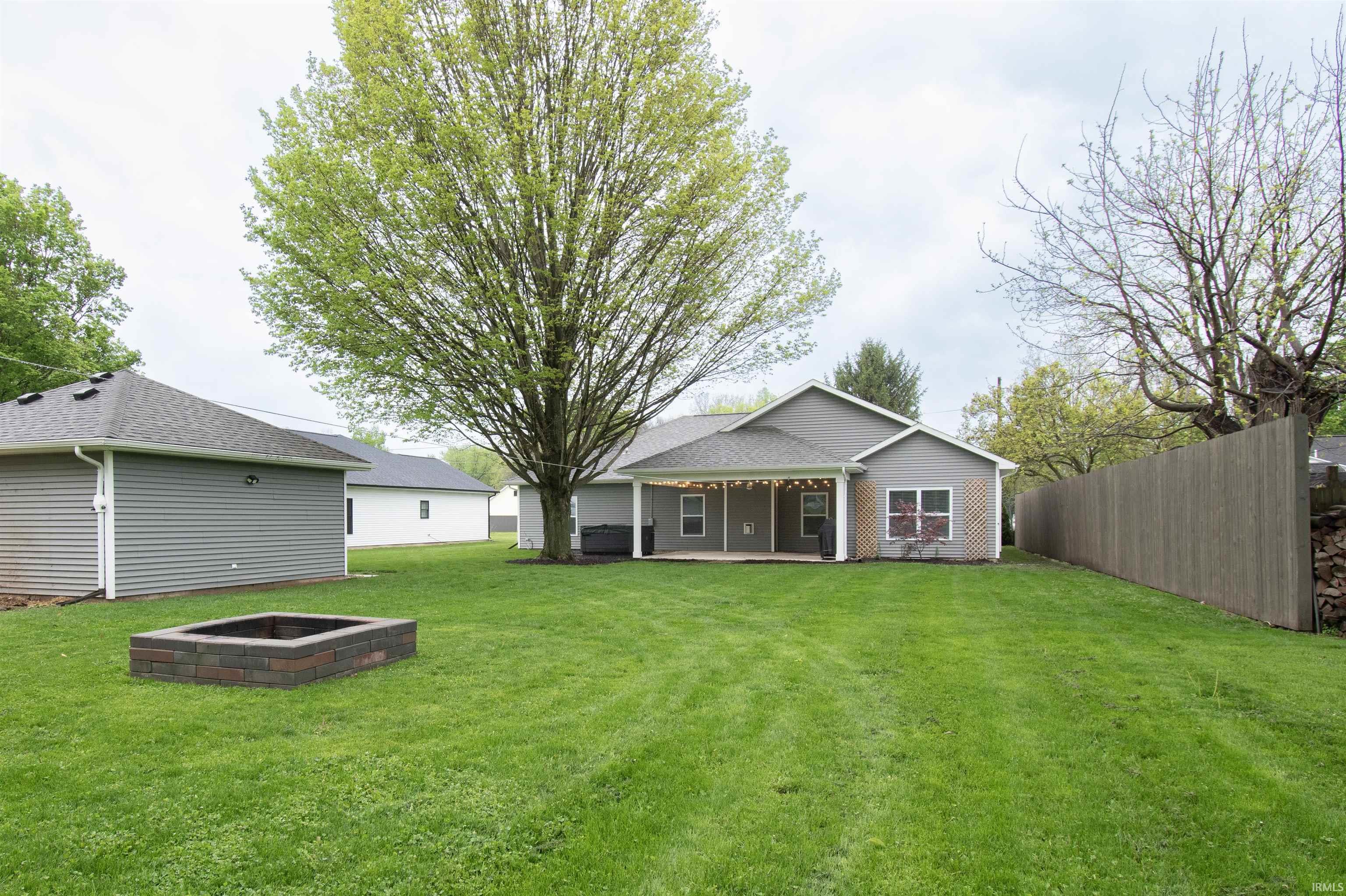 Expansive rear yard featuring a built-in stone fire pit, mature shade trees, and a wooden privacy fence