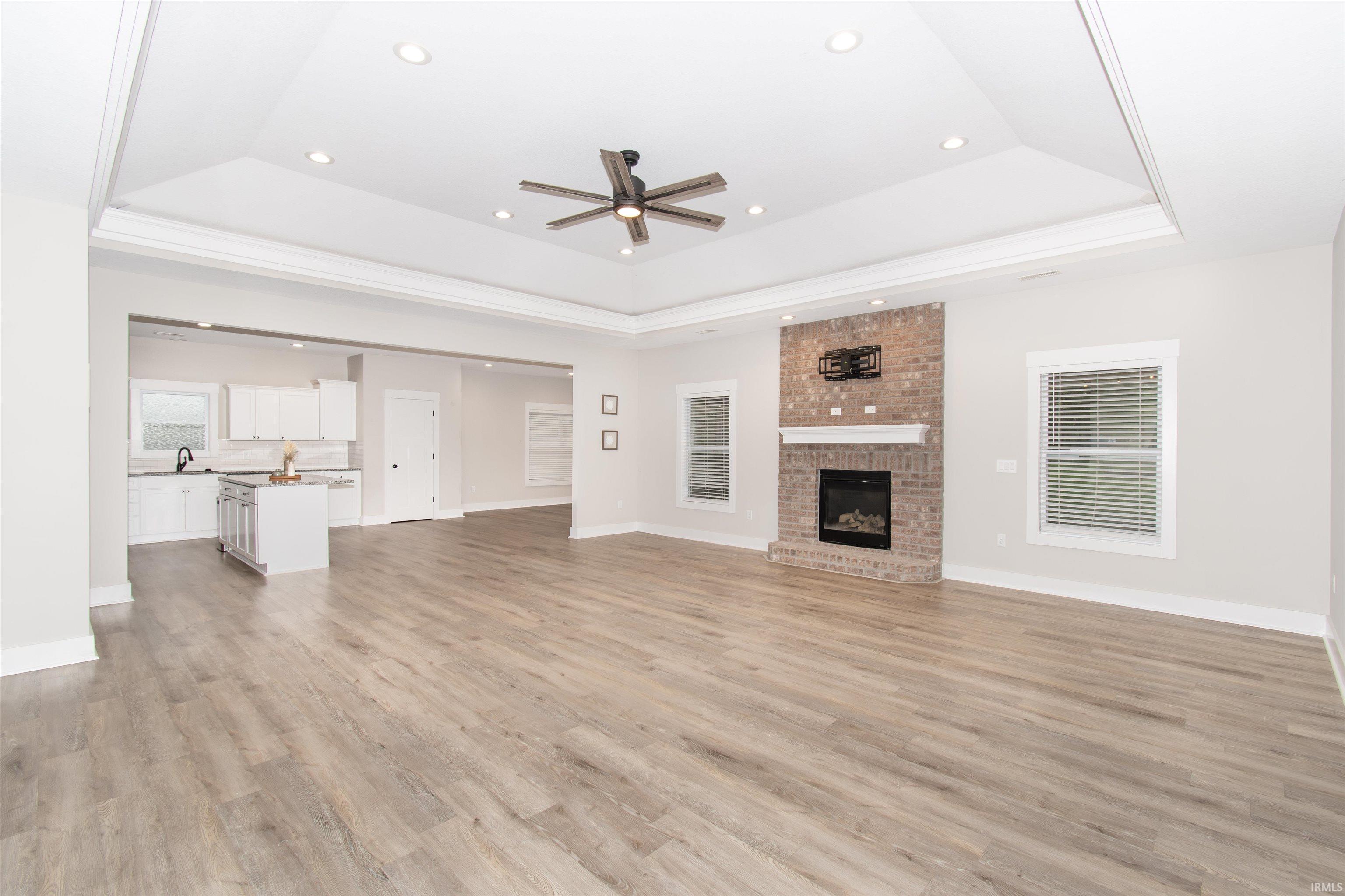 Spacious living area featuring wood-finish flooring, a brick fireplace with a white mantel, and a tray ceiling with recessed lighting and a ceiling fan