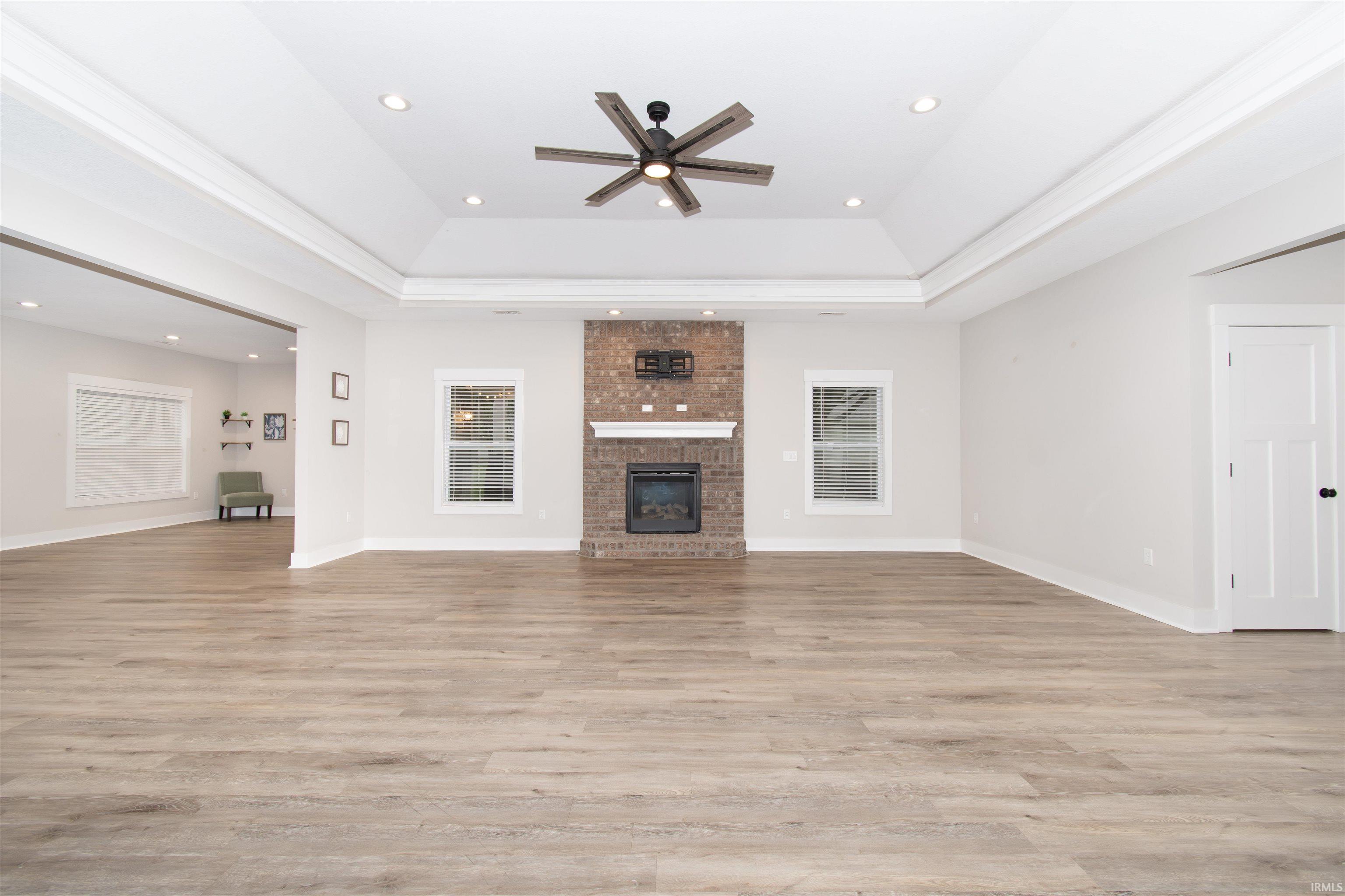 Spacious living area featuring wood-finish flooring, a brick-surround fireplace with a white mantel, and a tray ceiling with recessed lighting and a ceiling fan