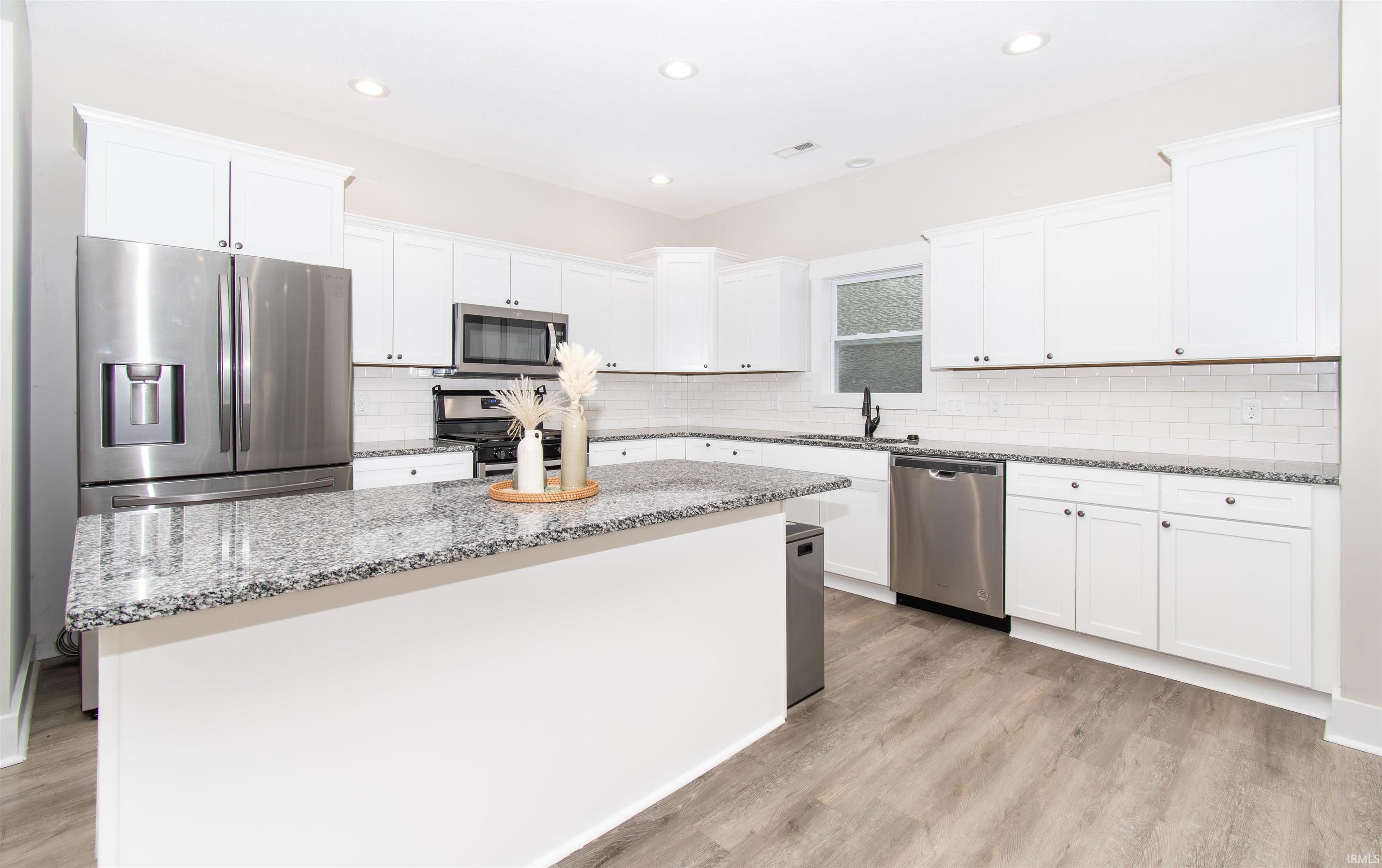 Spacious kitchen featuring white cabinetry, light granite countertops, stainless steel appliances, subway tile backsplash, and wood-finish flooring