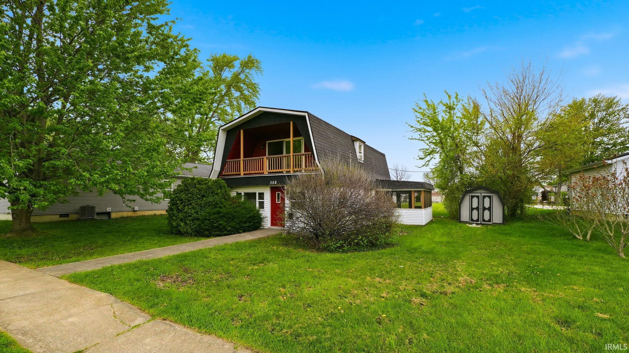 Gambrel roofline property featuring a second-story balcony with wood railing, a red entry door, a covered porch addition, and a detached storage shed