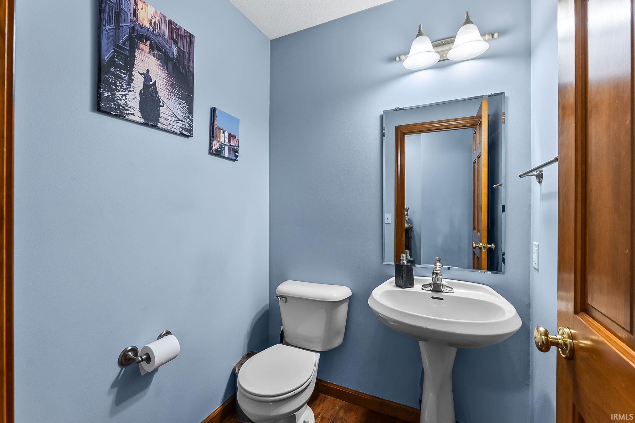 Powder room featuring a white pedestal sink, toilet, wood-finish flooring, and a mirrored wall fixture