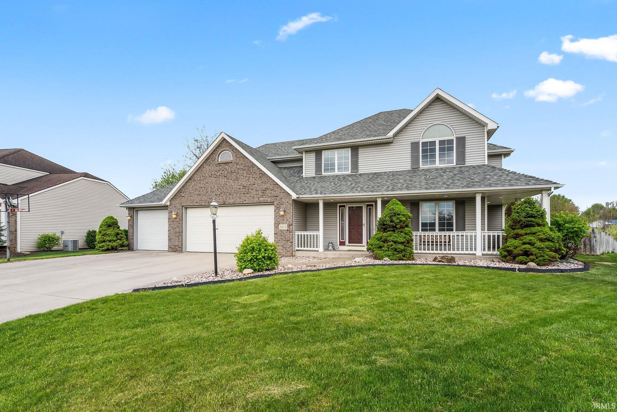 Two-story residence featuring a covered front porch with white railing, a grey siding and brick exterior, a two-car garage, an additional single-car garage, and a concrete driveway