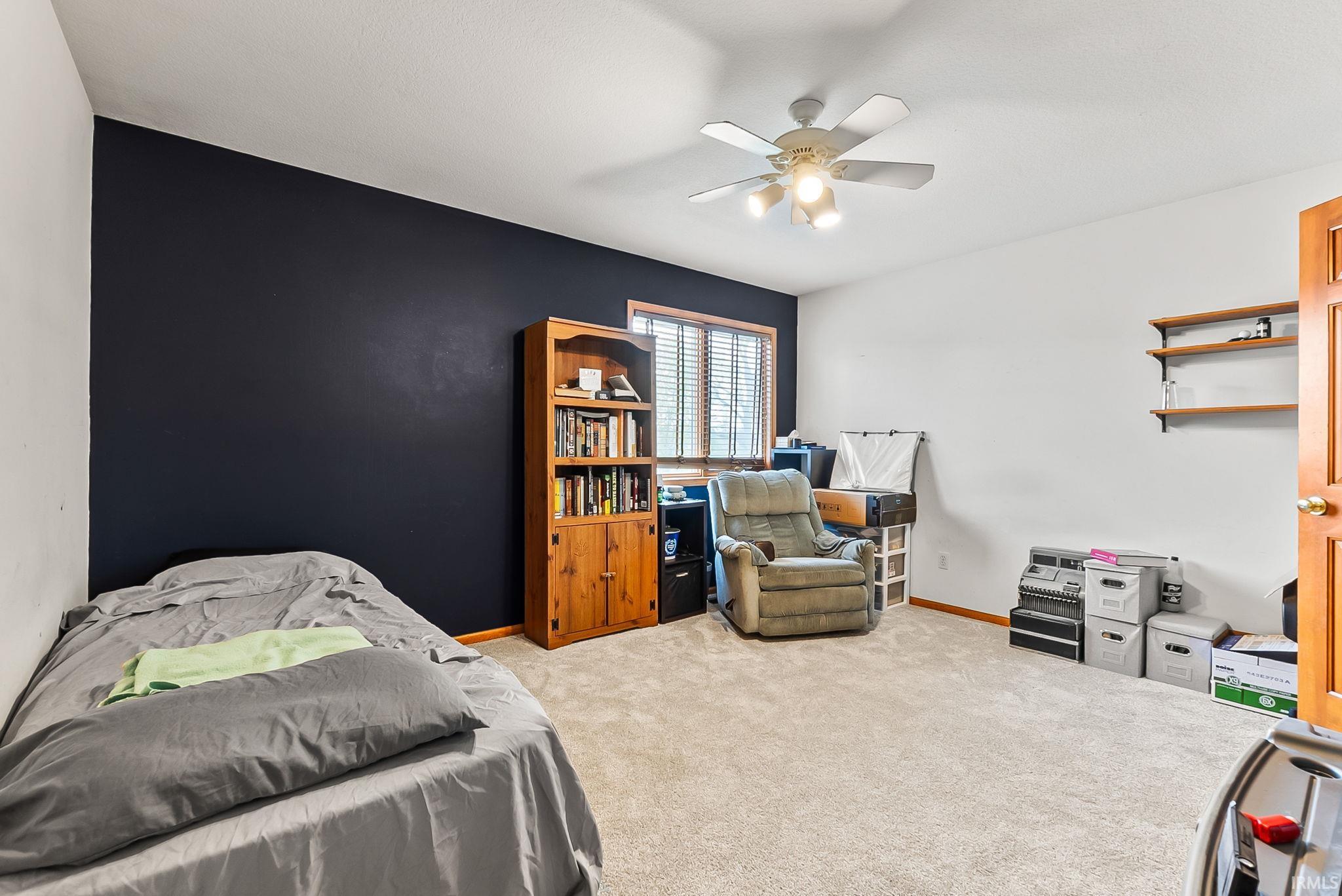 Well-proportioned room featuring a ceiling fan with integrated lighting, carpeted flooring, a window with blinds, and built-in wall shelving