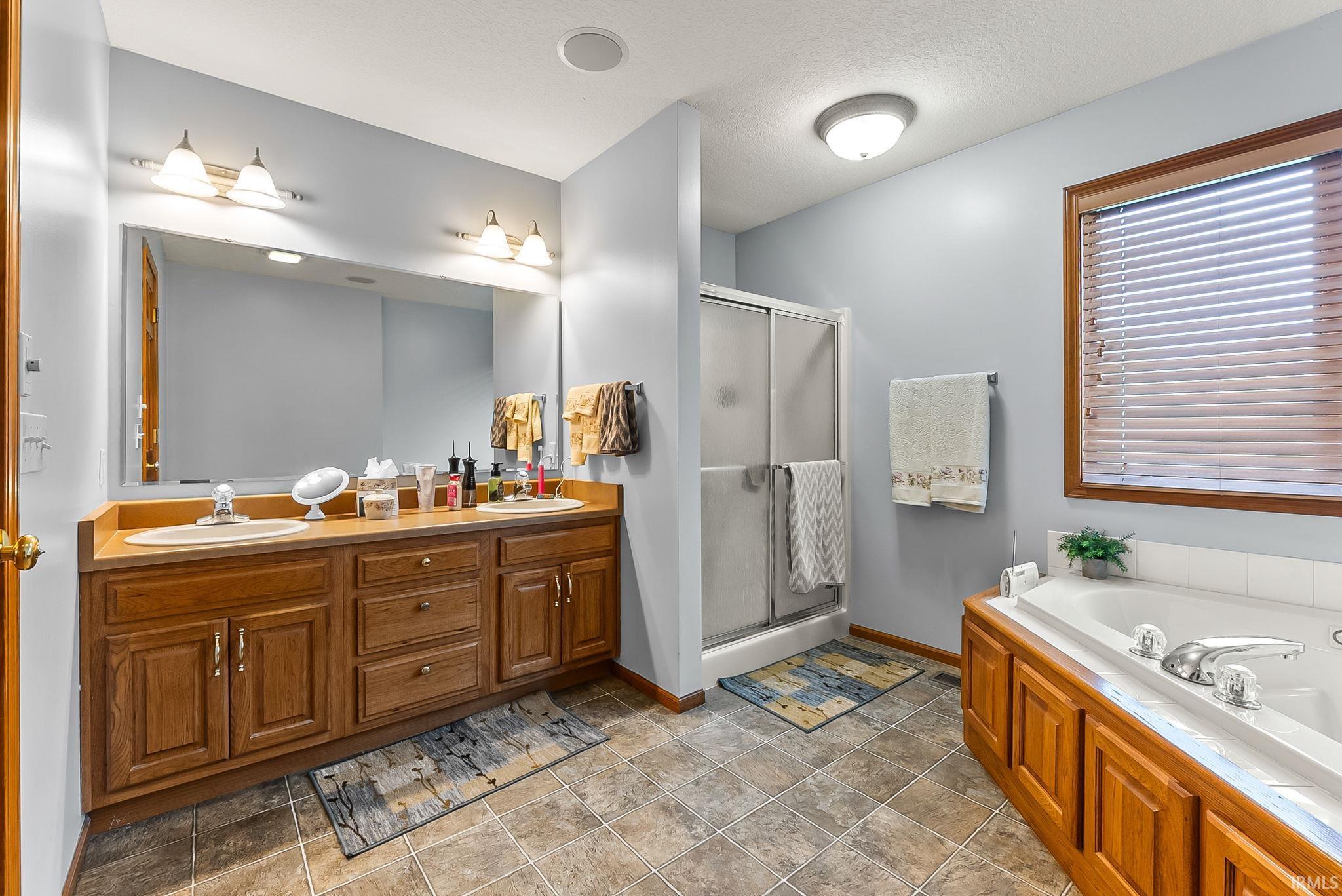 Bathroom featuring a dual-sink vanity with wood cabinetry, a built-in jetted tub, a separate glass-enclosed shower, and tiled flooring