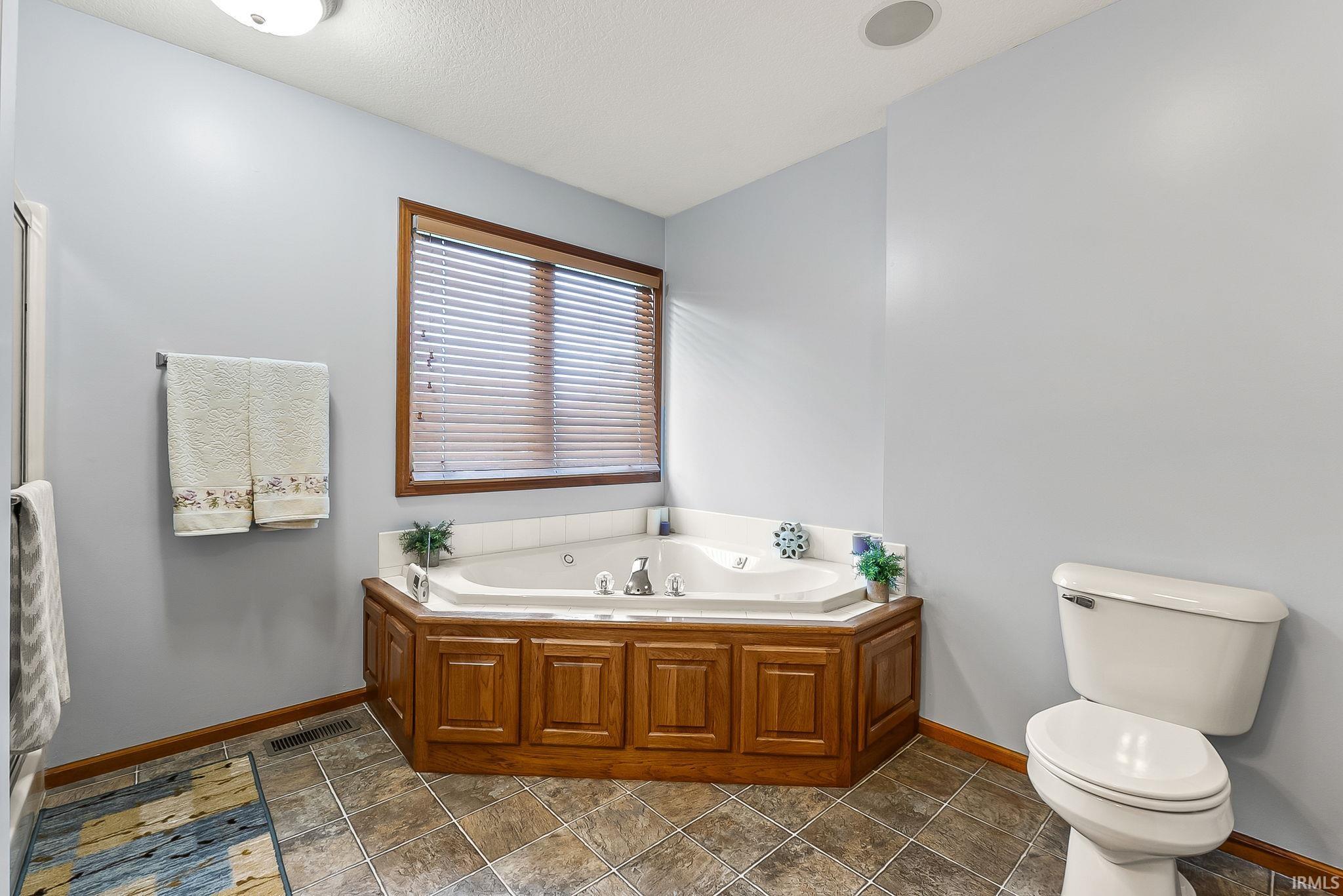 Corner jetted tub featuring a wood-finish surround and an adjacent window with blinds