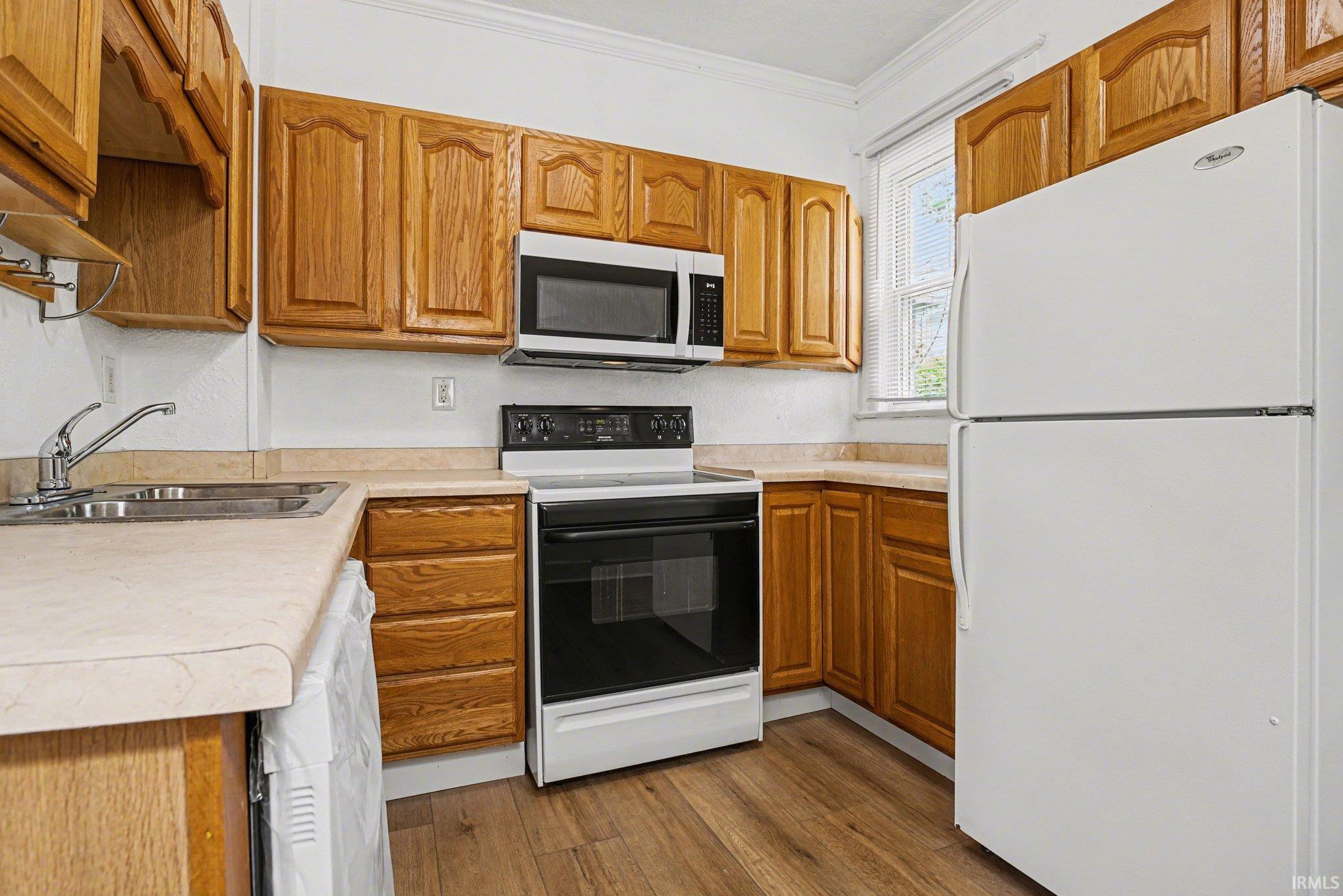 Kitchen featuring wood-finish cabinetry, light-toned countertops, a stainless steel double sink, a white electric range with a microwave above, and a white refrigerator